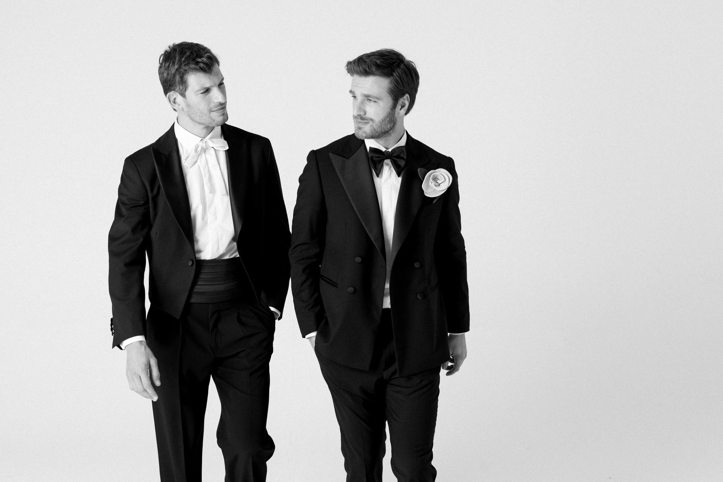 Due uomini eleganti in abiti da sera, uno con fiore sulla giacca, camminano e si guardano