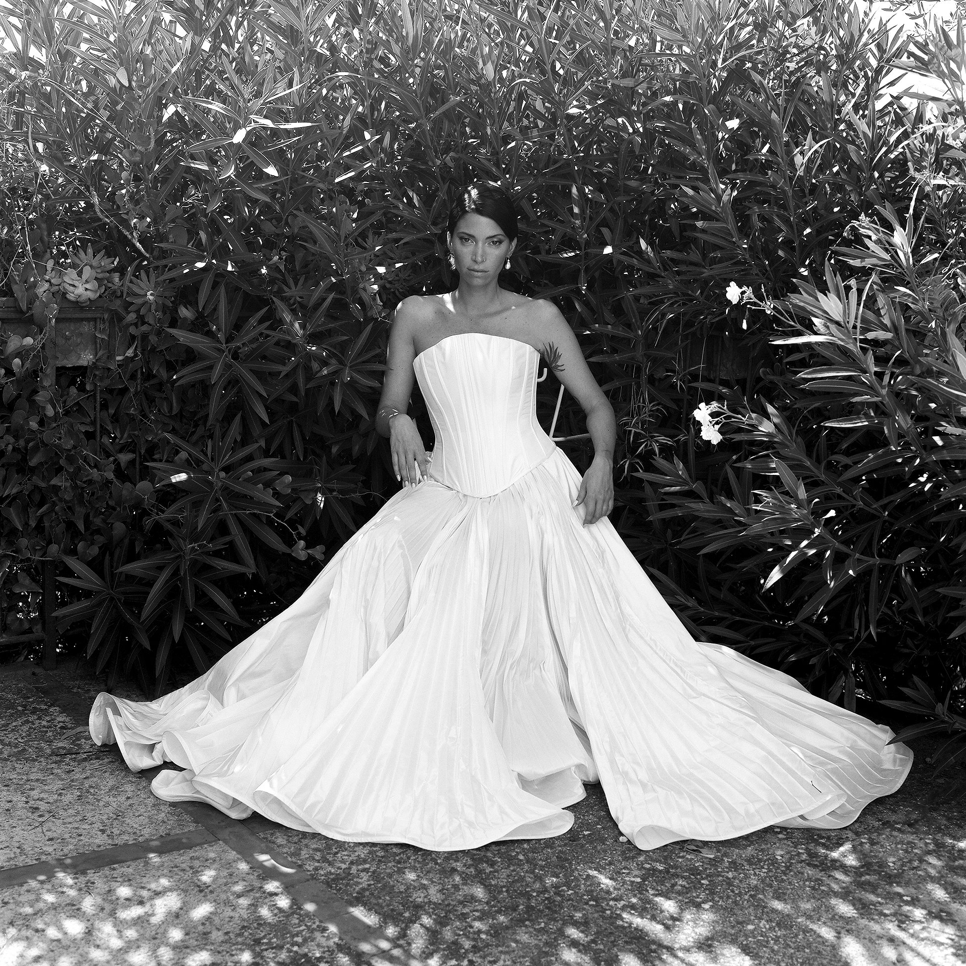 Donna in abito da sposa bianco con sfondo di cespugli e piante, in posa su pavimento.