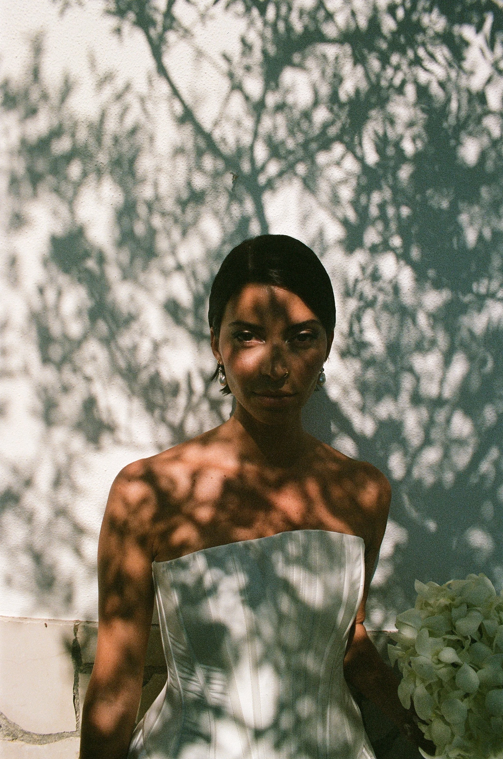Una donna indossa un vestito bianco strapless e si trova davanti a un muro con ombre di rami di alberi, creando un motivo di ombre sulla sua pelle e sullo sfondo.