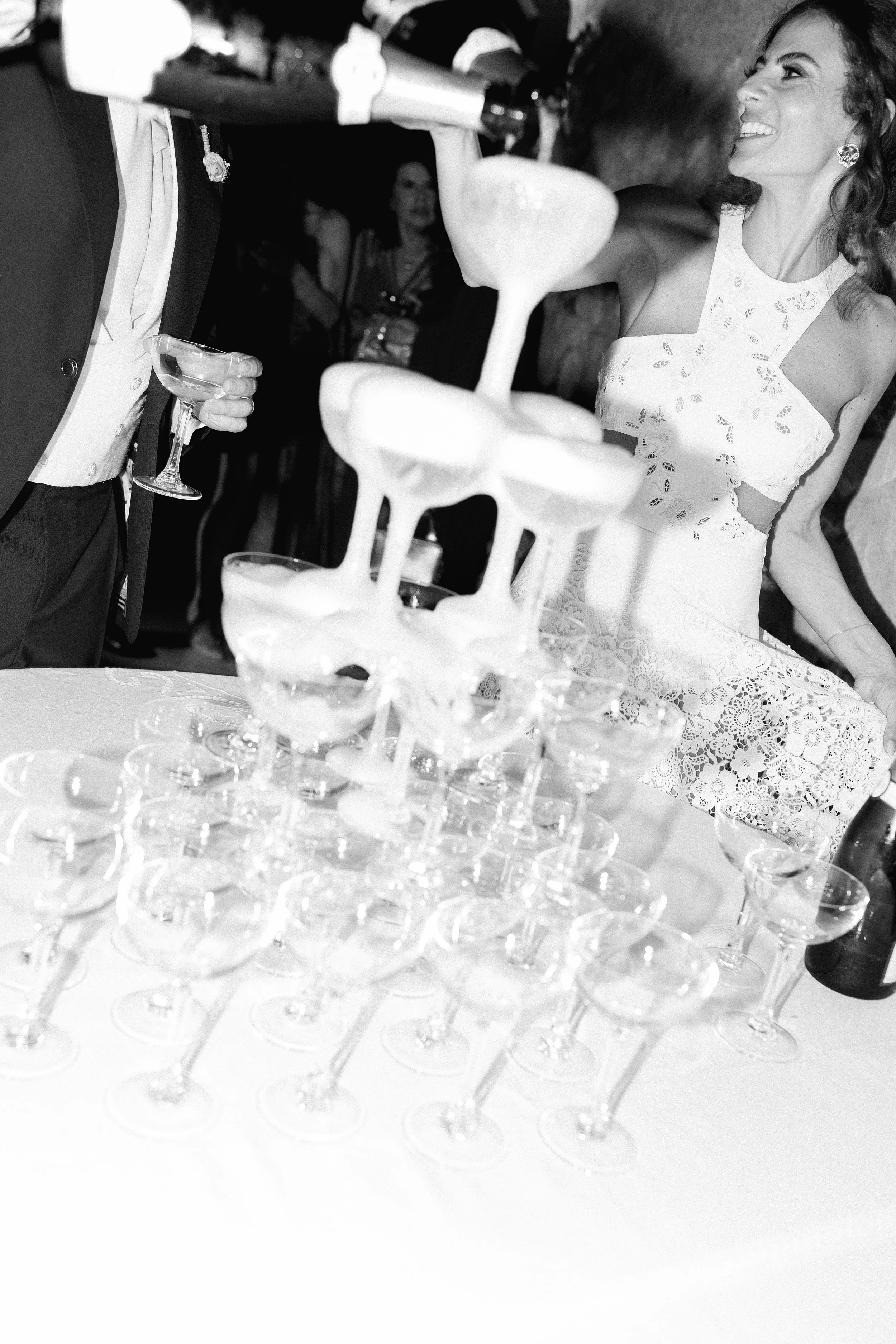 Torre di bicchieri di champagne su un tavolo durante una festa di matrimonio, con una donna in abito da sposa che sorride sullo sfondo.