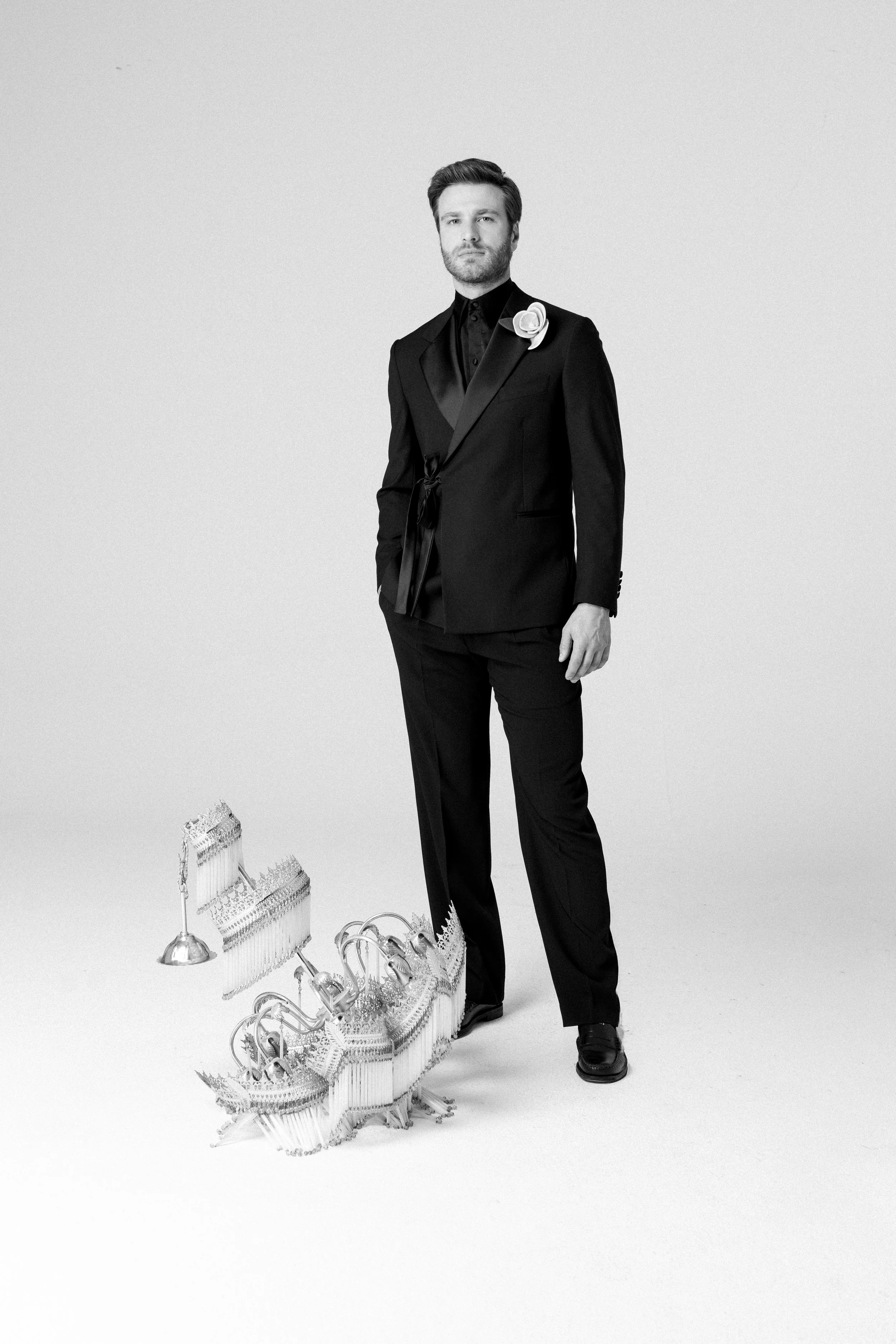 Un uomo in abito nero con boutonnière, che si erge accanto a un grande trofeo a forma di barca a vela, in un'immagine in bianco e nero.