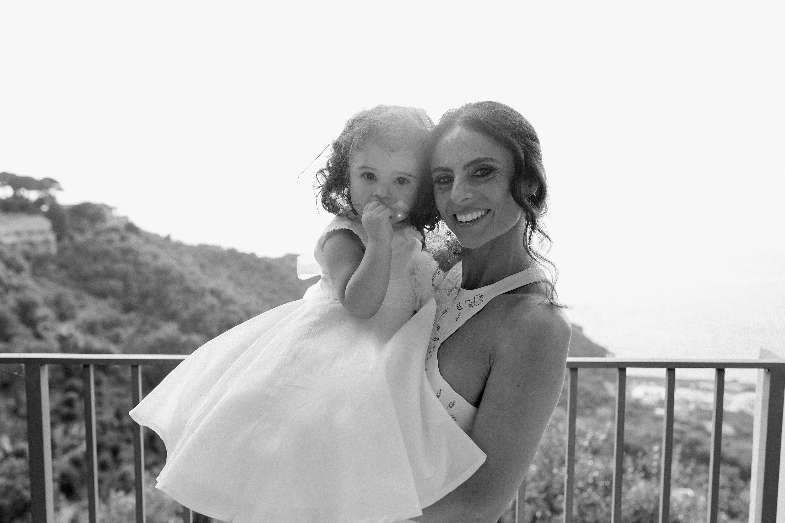 Una donna e una bambina sorridenti, la donna tiene la bambina in braccio, sullo sfondo un paesaggio collinare con alcuni edifici e un cielo chiaro.