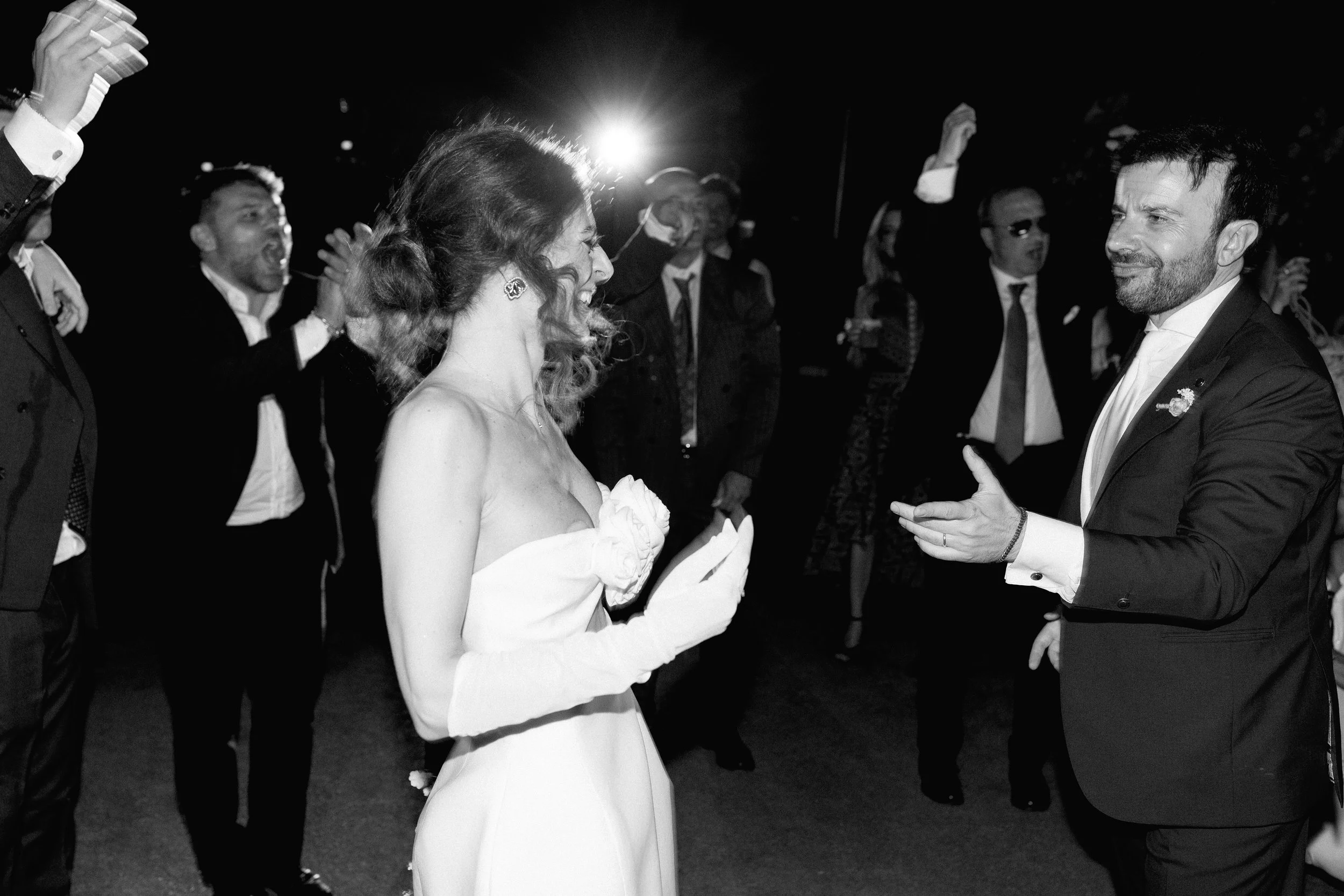 Una coppia di persone in abiti eleganti si guarda sorridendo durante un evento serale, circondata da altre persone in abiti formali.