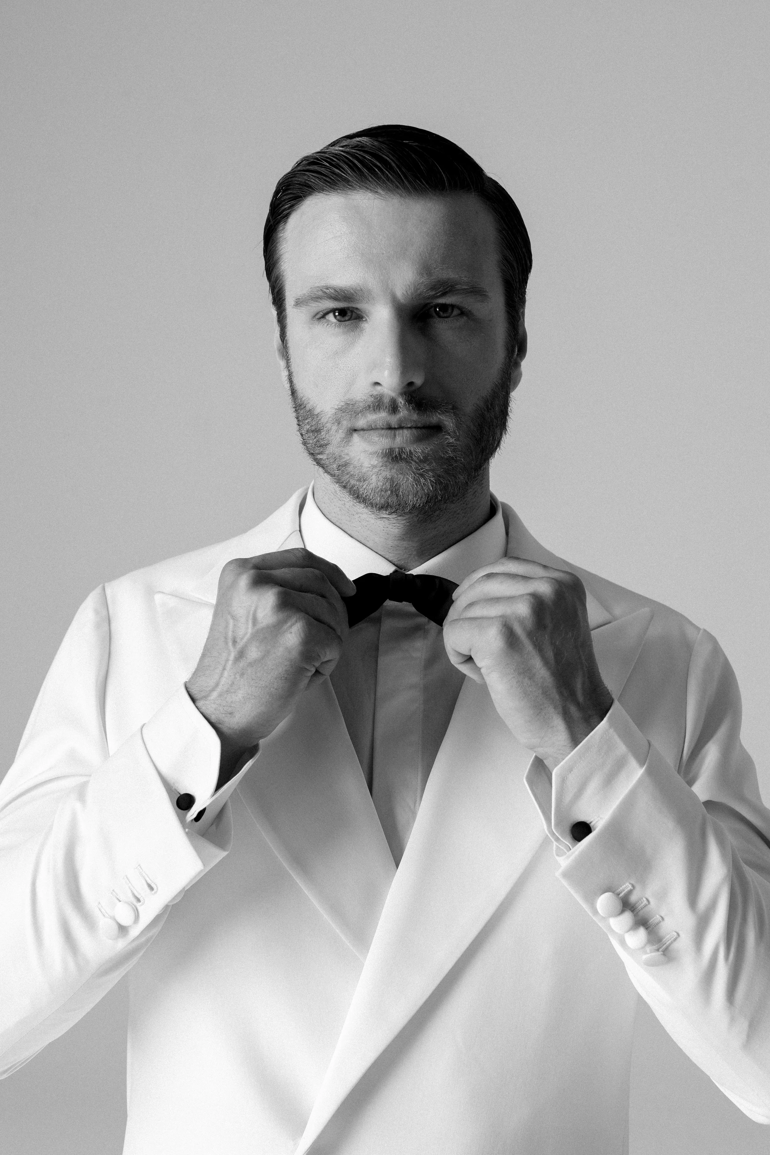 Uomo elegante con barba, in abito bianco, aggiustandosi un papillon su uno sfondo neutro.