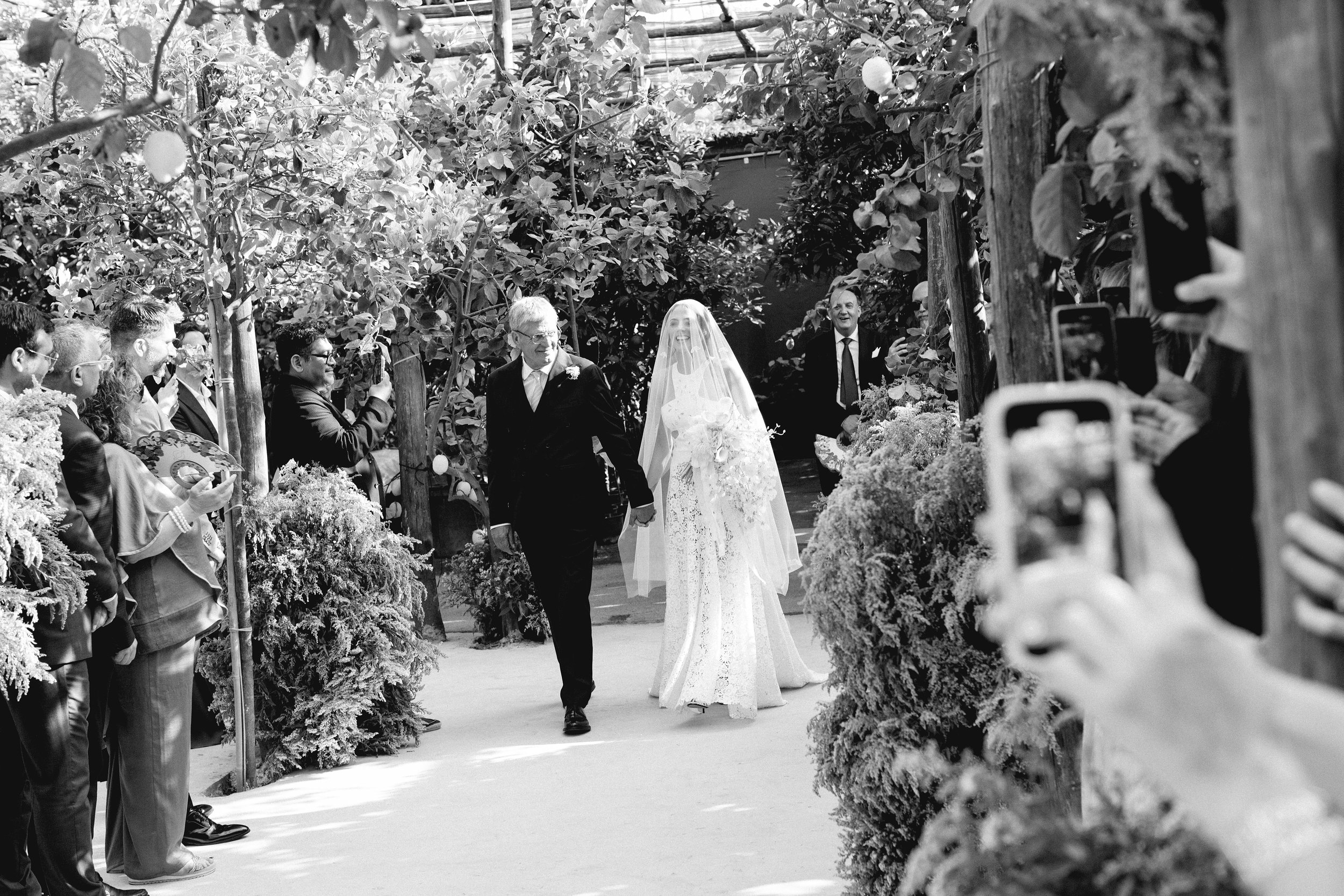 Una sposa in abito bianco e velo, accompagnata da un uomo anziano, cammina lungo un sentiero decorato con piante e fiori, circondata da persone che la scattano con lo smartphone durante un matrimonio.