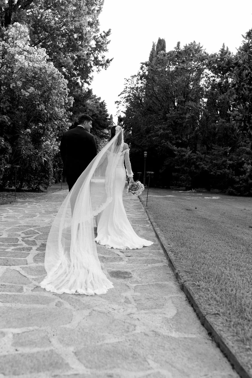Una coppia di sposi cammina lungo un sentiero in un parco, indossando abiti da matrimonio, con la sposa che tiene un bouquet di fiori.