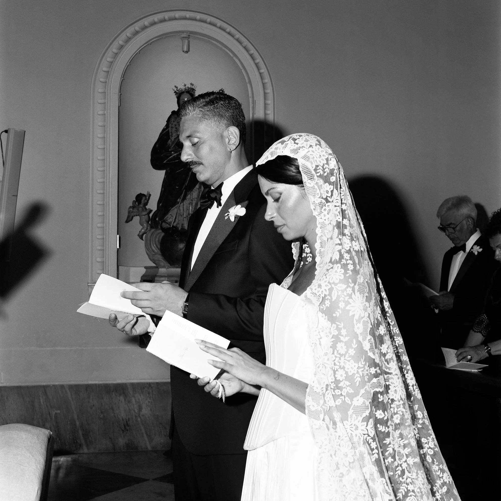 Coppia di sposi durante il matrimonio, che leggono qualcosa, con altri presenti che fanno lo stesso.