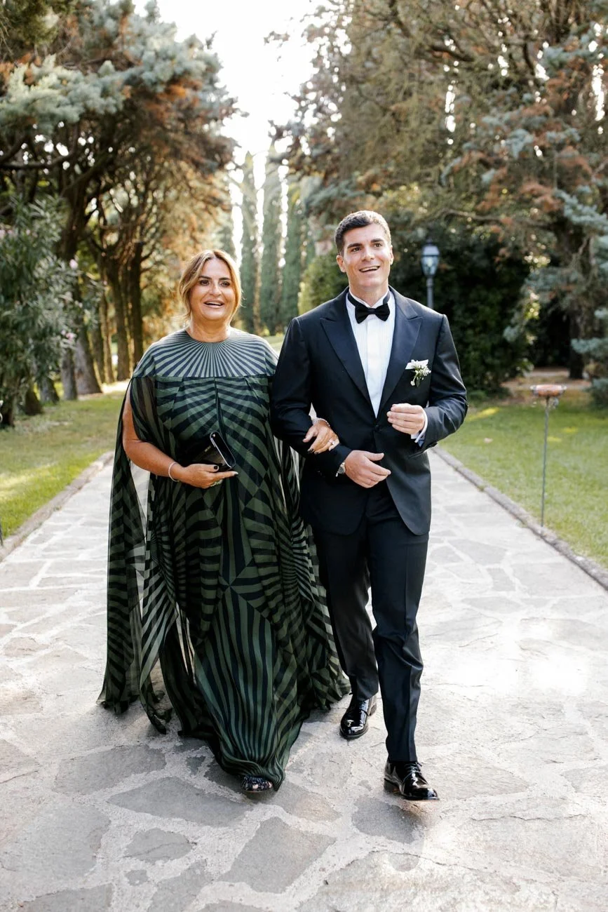 Una coppia ben vestita, l'uomo in smoking e la donna con un abito lungo in stile elegante, camminano insieme in un giardino. Sembrano essere in occasione di un matrimonio, circondati da alberi e vegetazione.