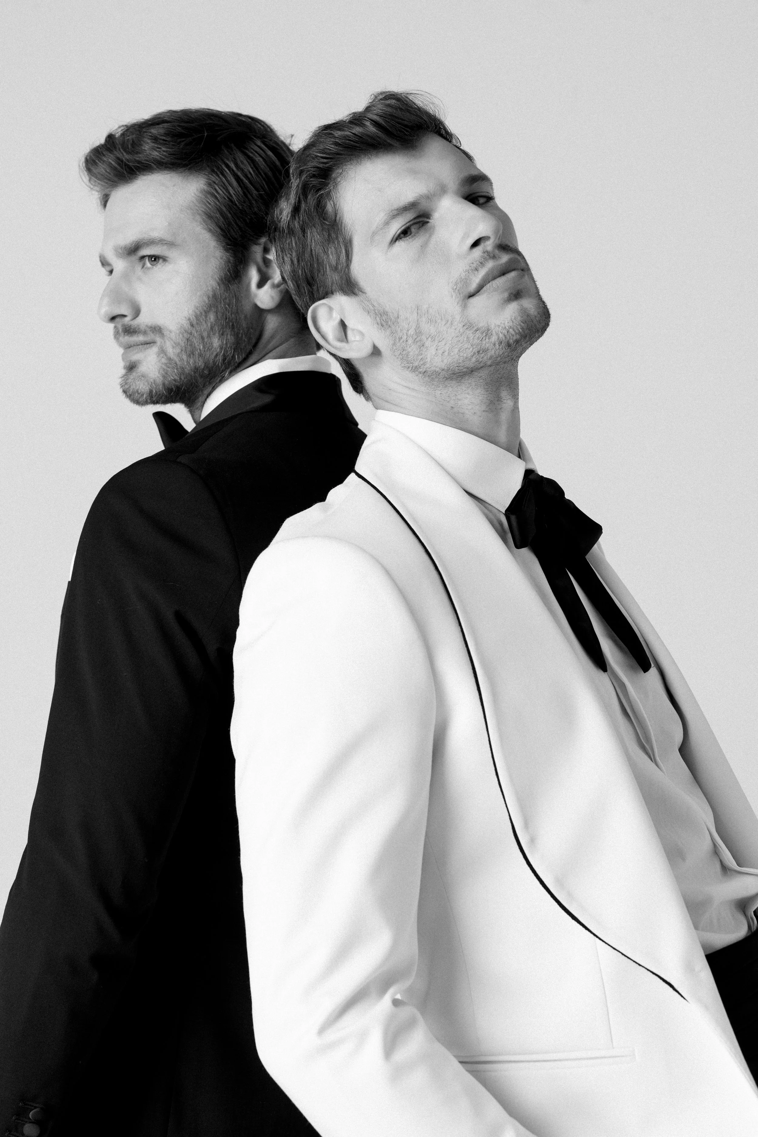 Due uomini eleganti in abiti da sera, uno con giacca bianca e cravatta nera, l'altro con giacca nera e cravatta bianca, posano uno dietro l'altro, in uno stile di moda sofisticata.