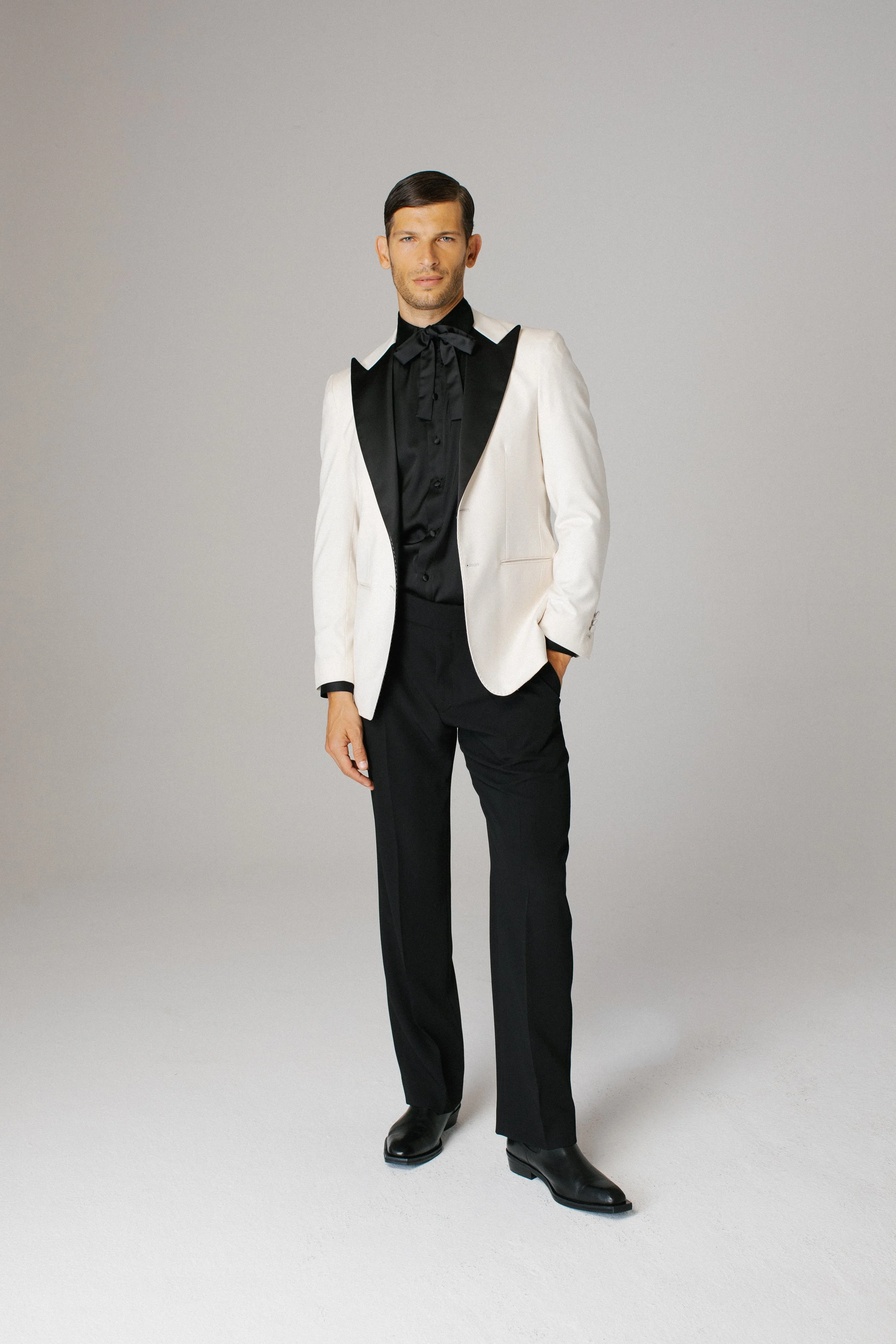 Uomo elegante con giacca bianca e camicia nera in piedi su sfondo neutro.