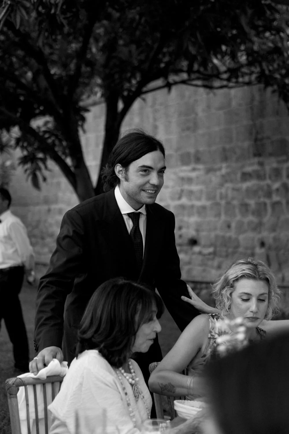 Foto in bianco e nero di una riunione tra donne sedute a un tavolo e un uomo in giacca e cravatta che si avvicina e sorride, all'aperto con un muro di pietra e un albero sullo sfondo.