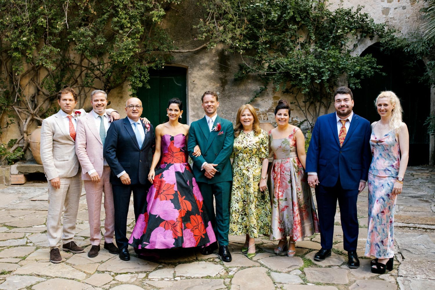 Gruppo di persone sorridenti, vestite elegantemente, in posa all'aperto su strada di pietra, con muri ricoperti di piante verdi sullo sfondo, durante un evento speciale o un matrimonio.