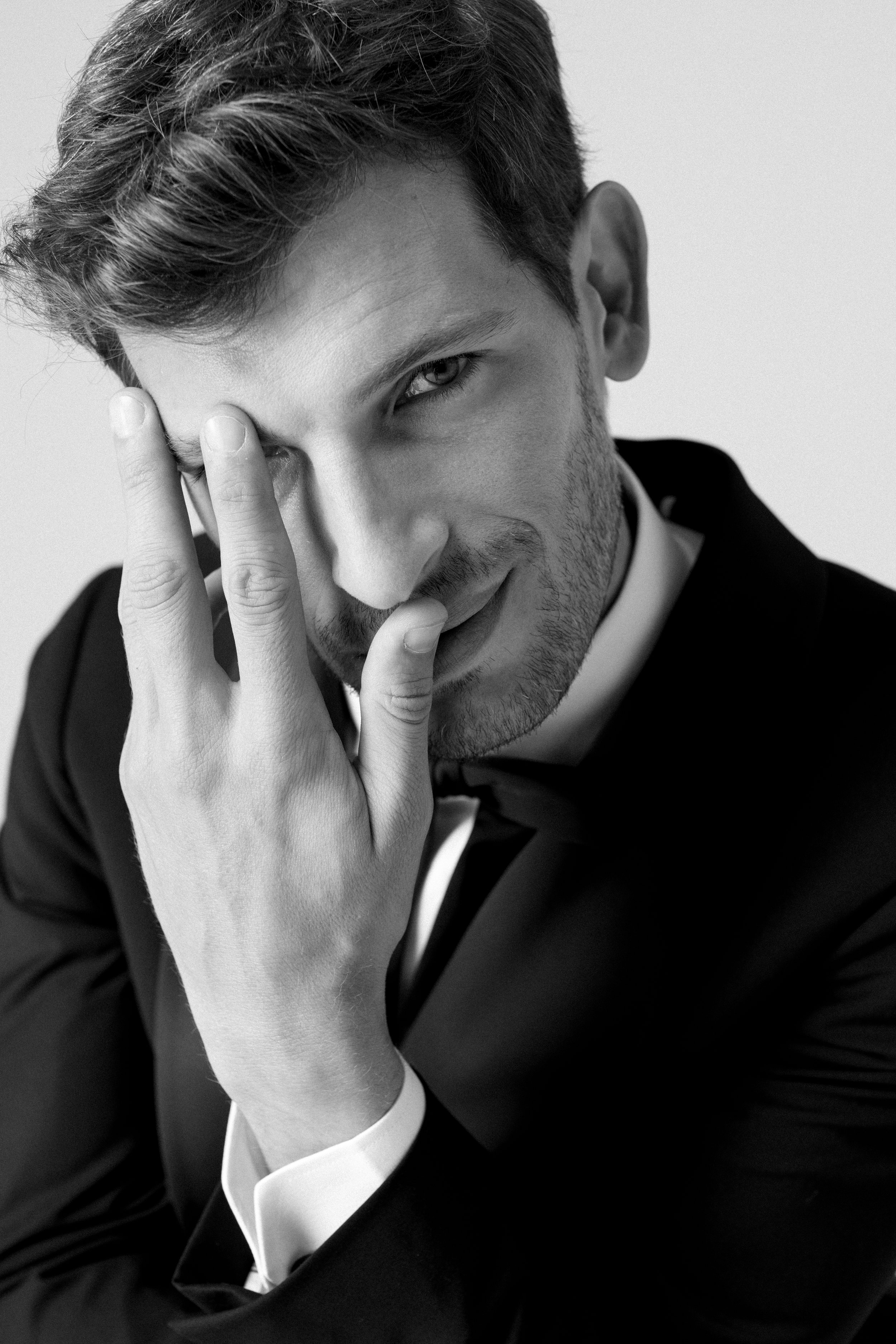 Ritratto di un uomo giovane che indossa un abito formale con un sorriso ambiguo, toccandosi il viso con una mano.