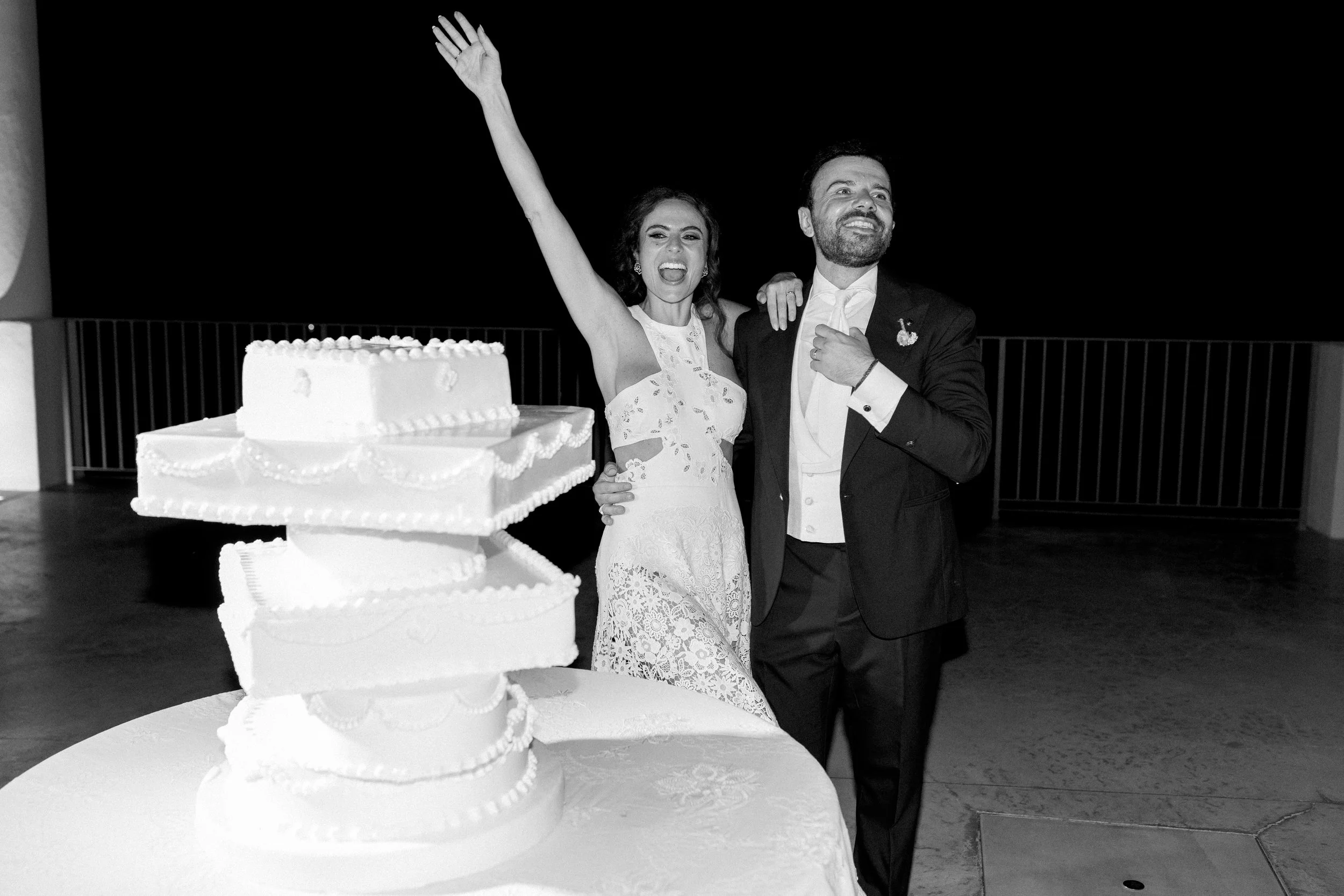 Una coppia di sposi al loro matrimonio con un grande torta nuziale davanti, sorridenti e felici.