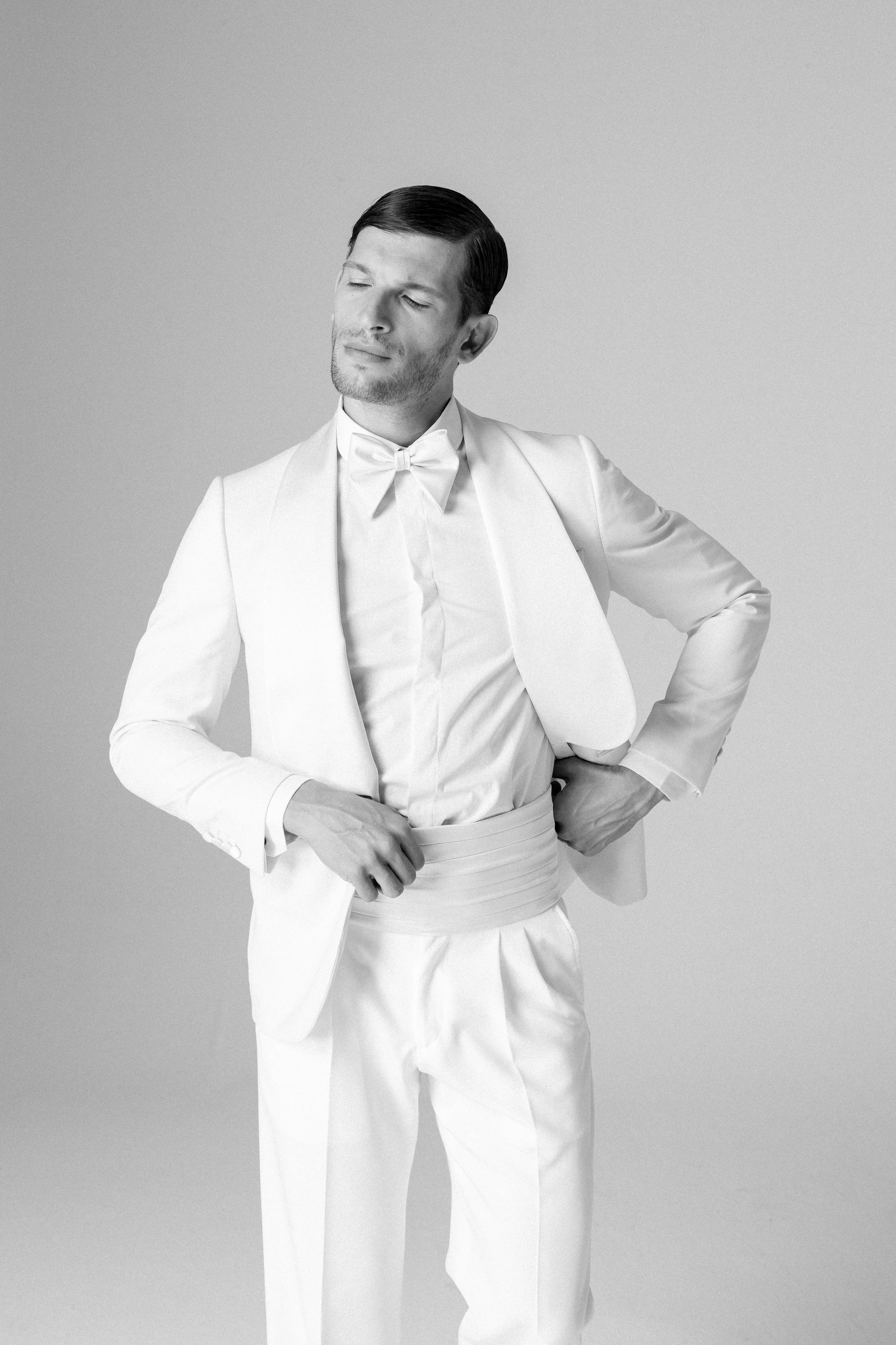 Un uomo vestito elegantemente in bianco, con giacca, pantaloni, camicia e fiocco, si trova contro uno sfondo semplice, con un'espressione pensierosa.