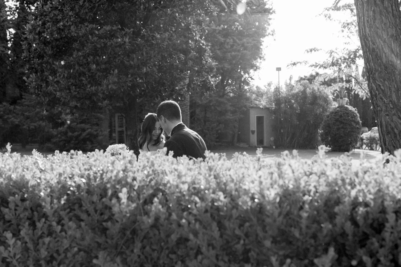 Una coppia si incontra in un parco ombreggiato, circondata da alberi e cespugli, con il sole che brilla dietro di loro.