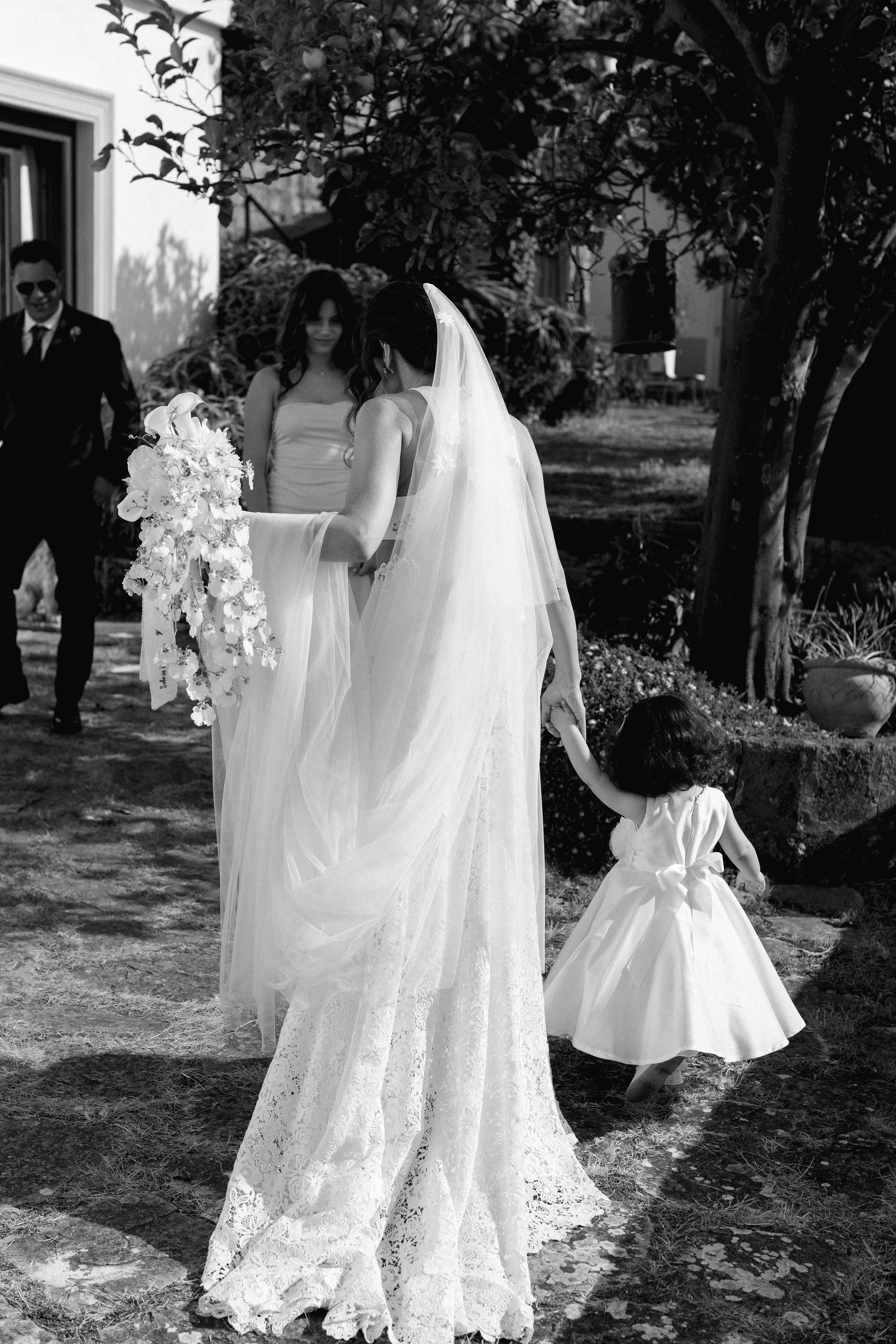 Una donna in abito da sposa tiene la mano di una bambina in un giardino, con altri adulti in abiti eleganti sullo sfondo.