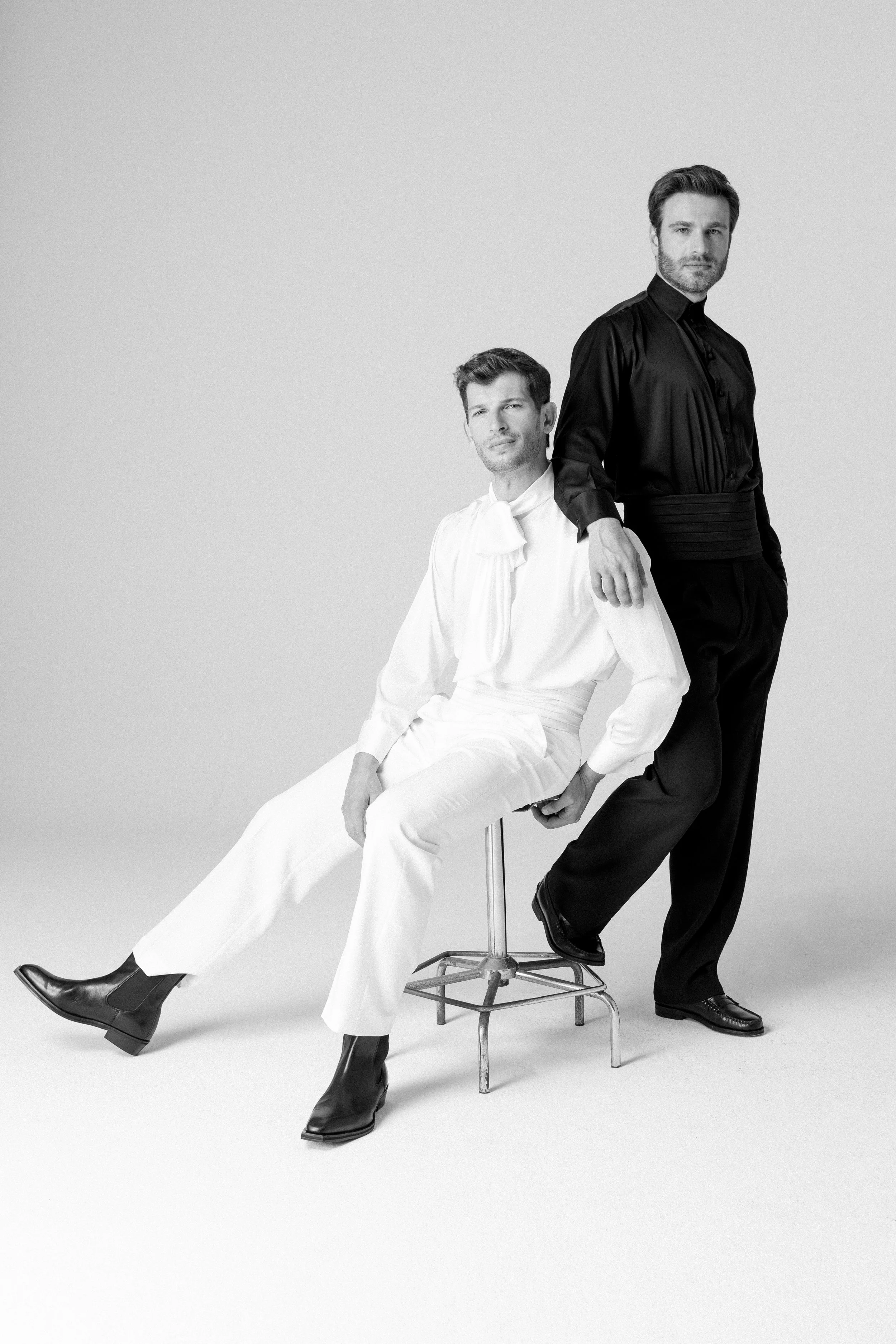 Due uomini vestiti elegantemente, uno seduto in abiti bianchi e uno in piedi in abiti neri, in una foto in bianco e nero.