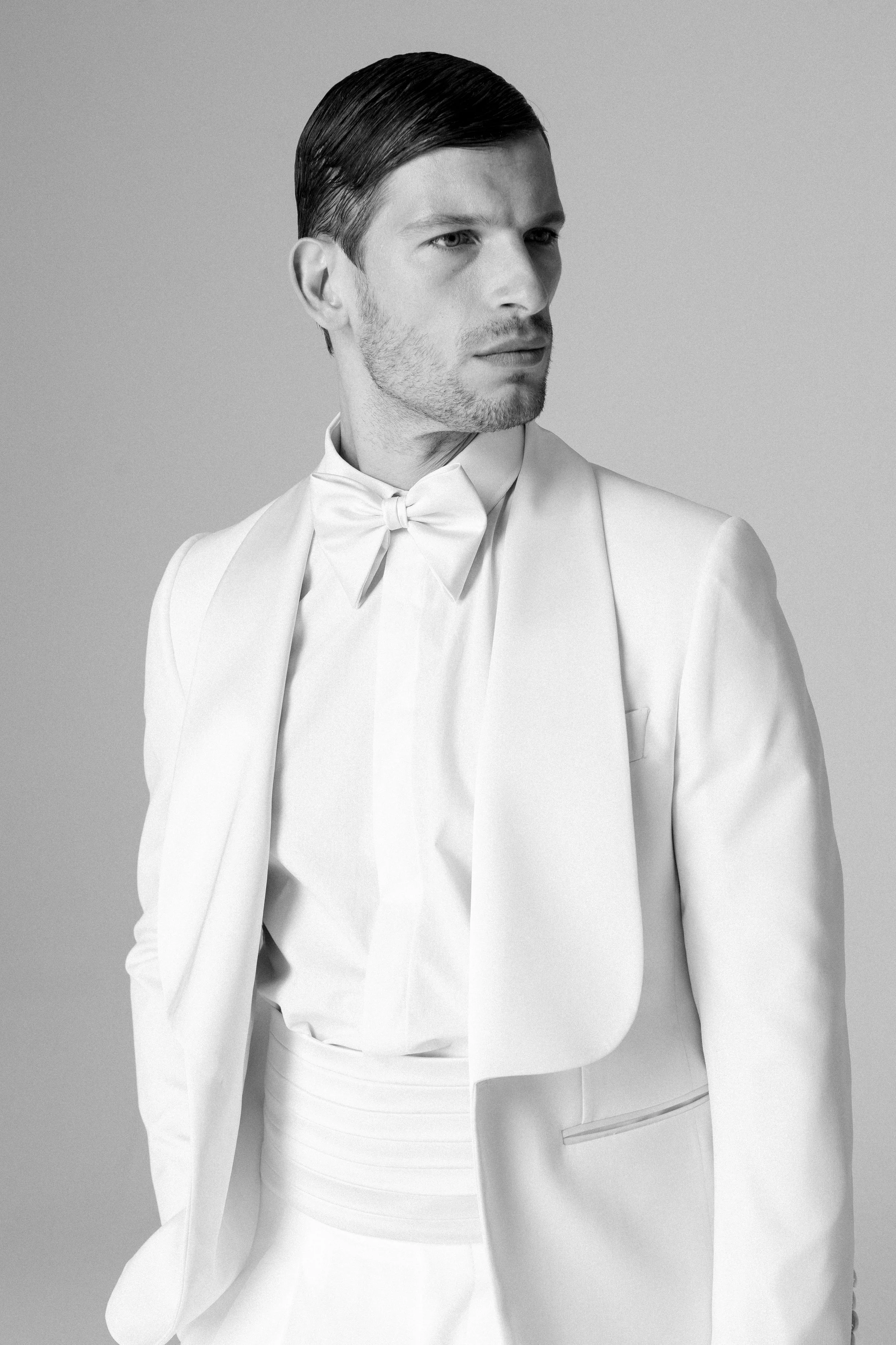 Uomo elegante in abito bianco con papillon, posa in un ambiente neutro, fotografia in bianco e nero.