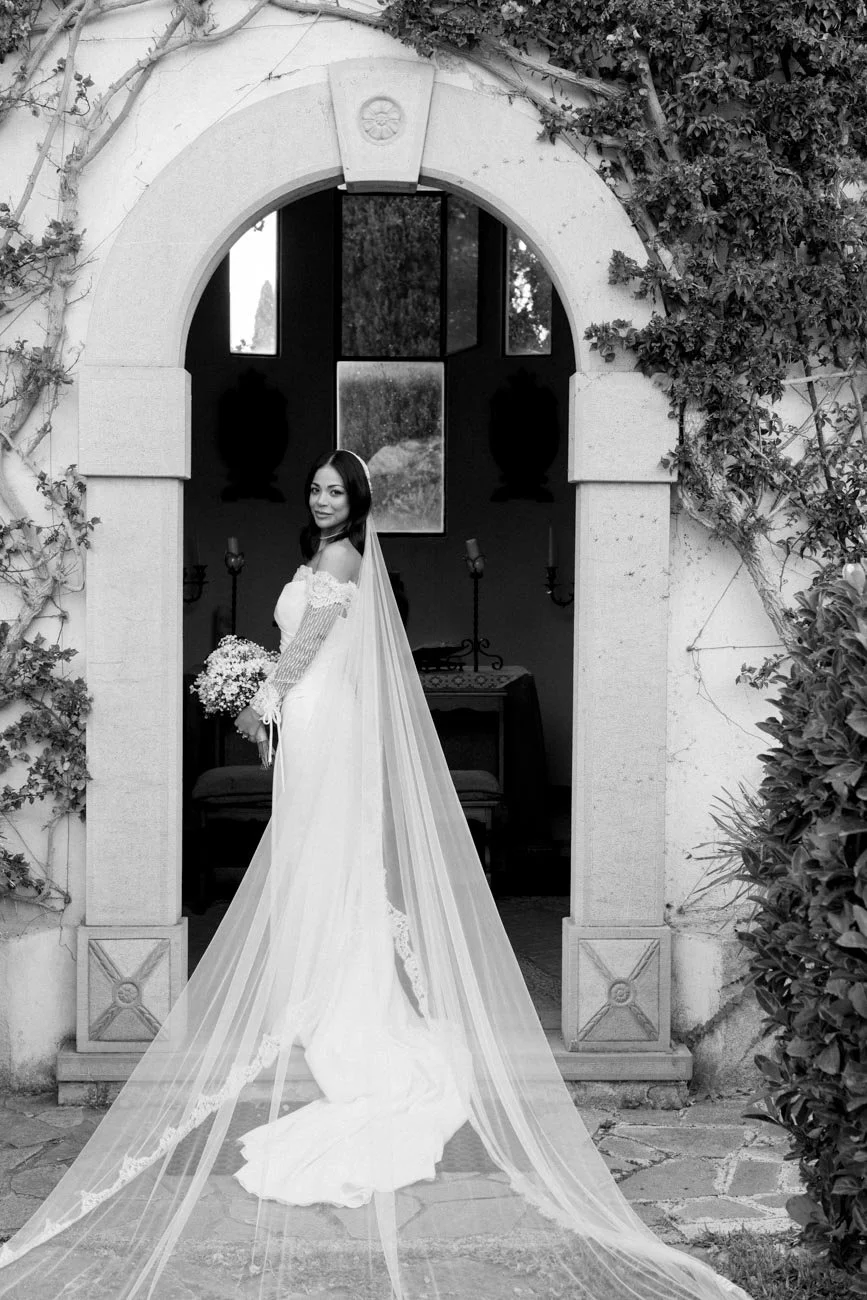 Una sposa in abito da cerimonia con un lungo velo bianco si trova davanti a un arco di pietra, circondato da piante.