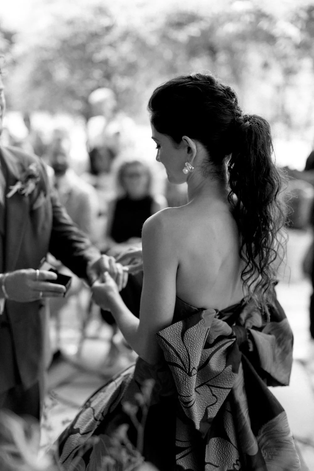 Donna con capelli ricci, indossa orecchini pendenti e un abito senza spalle con una grande sciarpa o wrap con motivo floreale, in un evento formale o cerimonia, in bianco e nero.