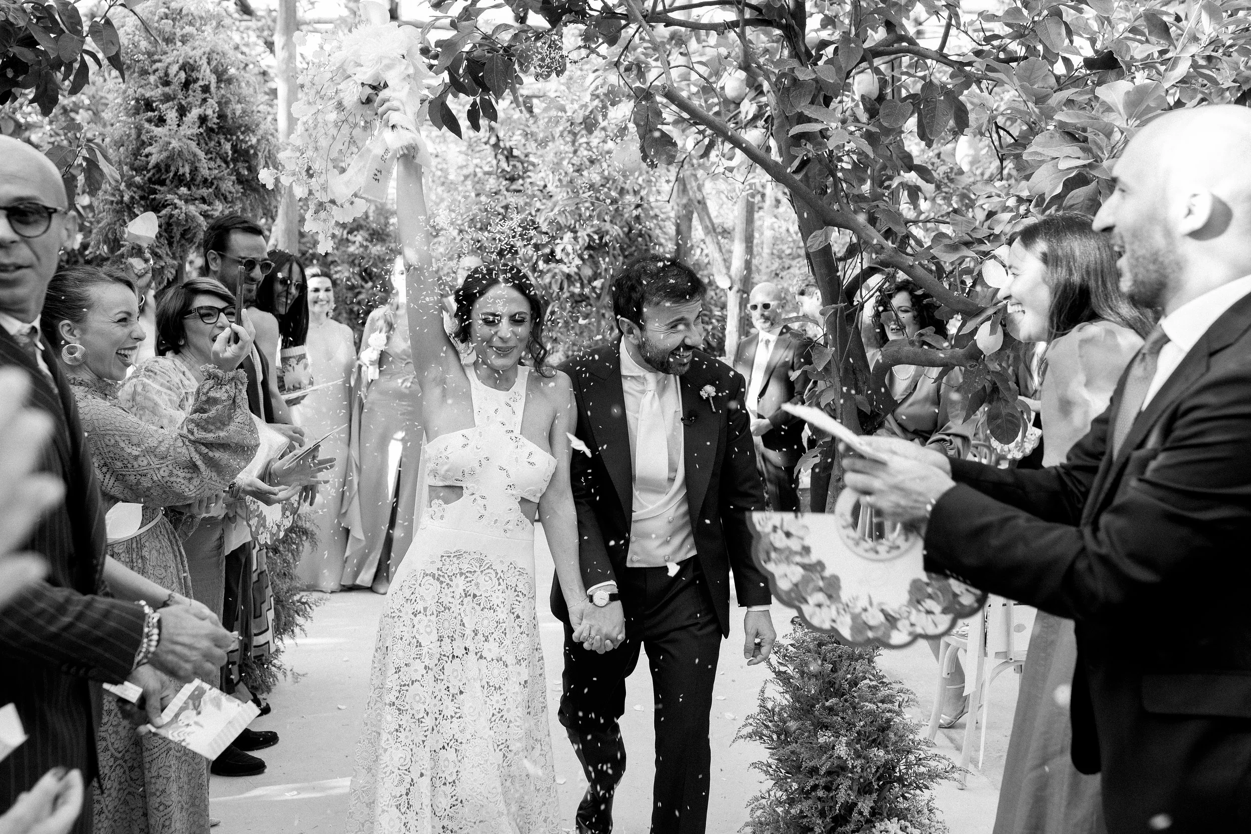 Una coppia di sposi felici che si tiene per mano, cammina sotto un arco di fiori mentre gli invitati li celebrano e lanciano petali o coriandoli, in un matrimonio.