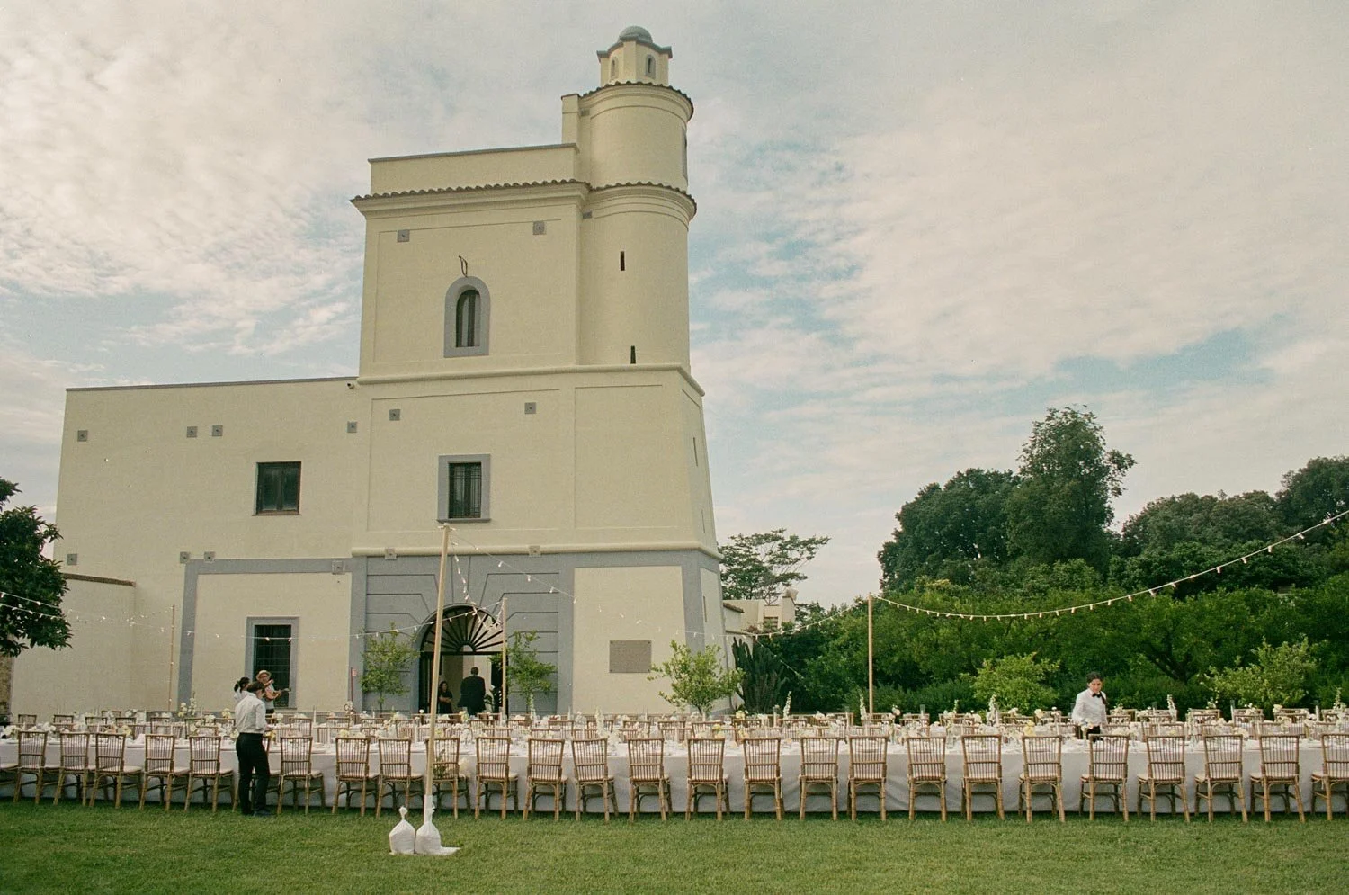 Un edificio bianco con torre, decorato per un evento all'aperto con tavoli e sedie, alcune persone che preparano l'evento e alberi sullo sfondo.