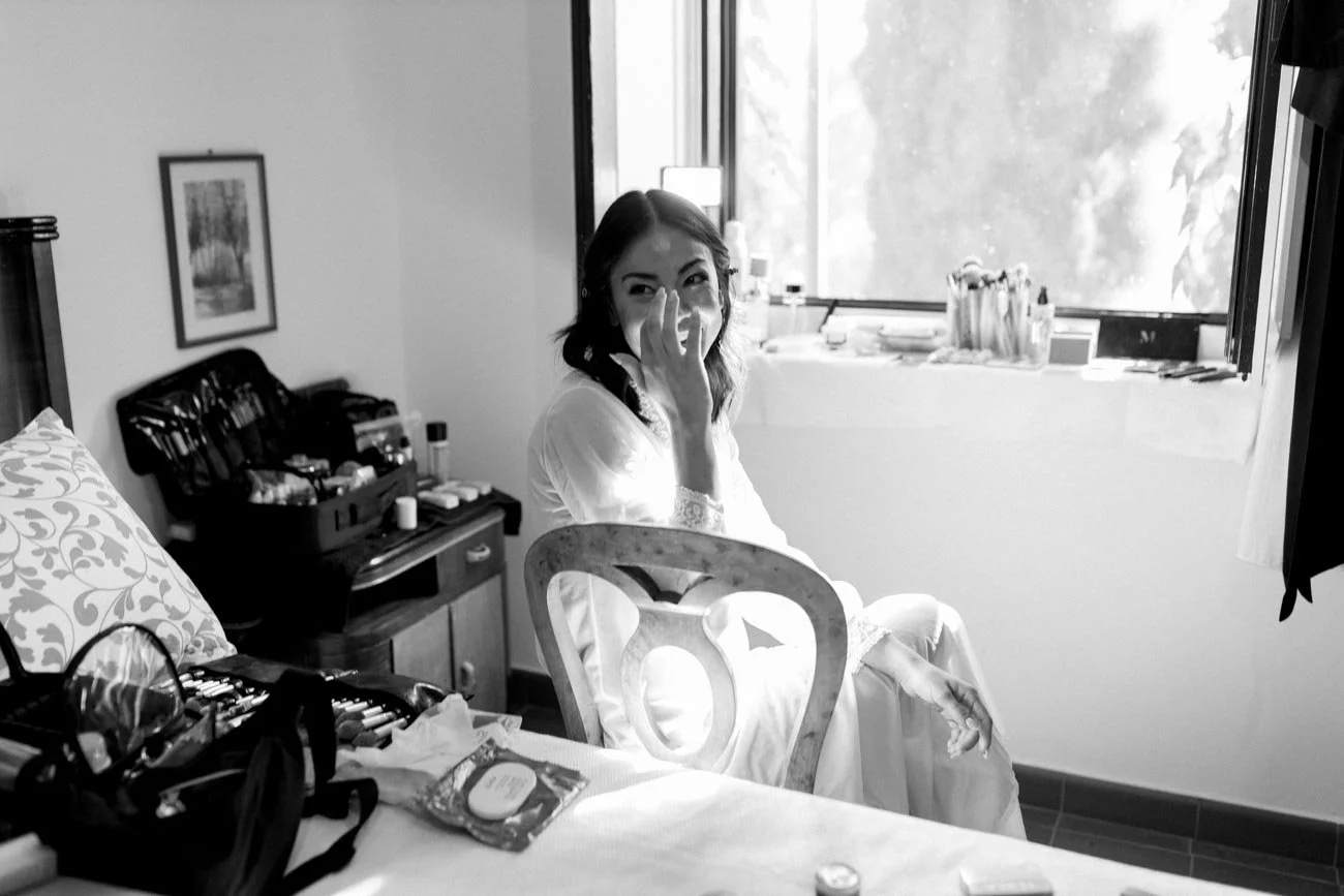 Donna sorridente seduta vicino a una finestra, mentre si sistema il volto, con vari oggetti di trucco e una stanza in stile classico sullo sfondo.