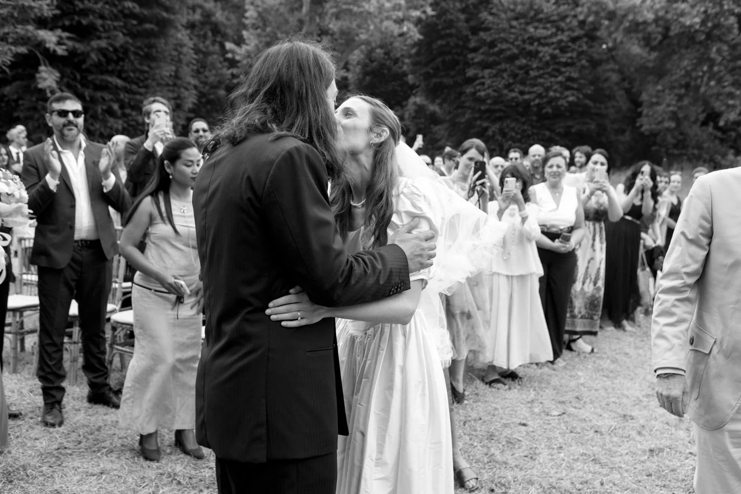 Due persone si baciano durante un matrimonio all'aperto, circondate da amici e familiari che osservano e scattano foto.