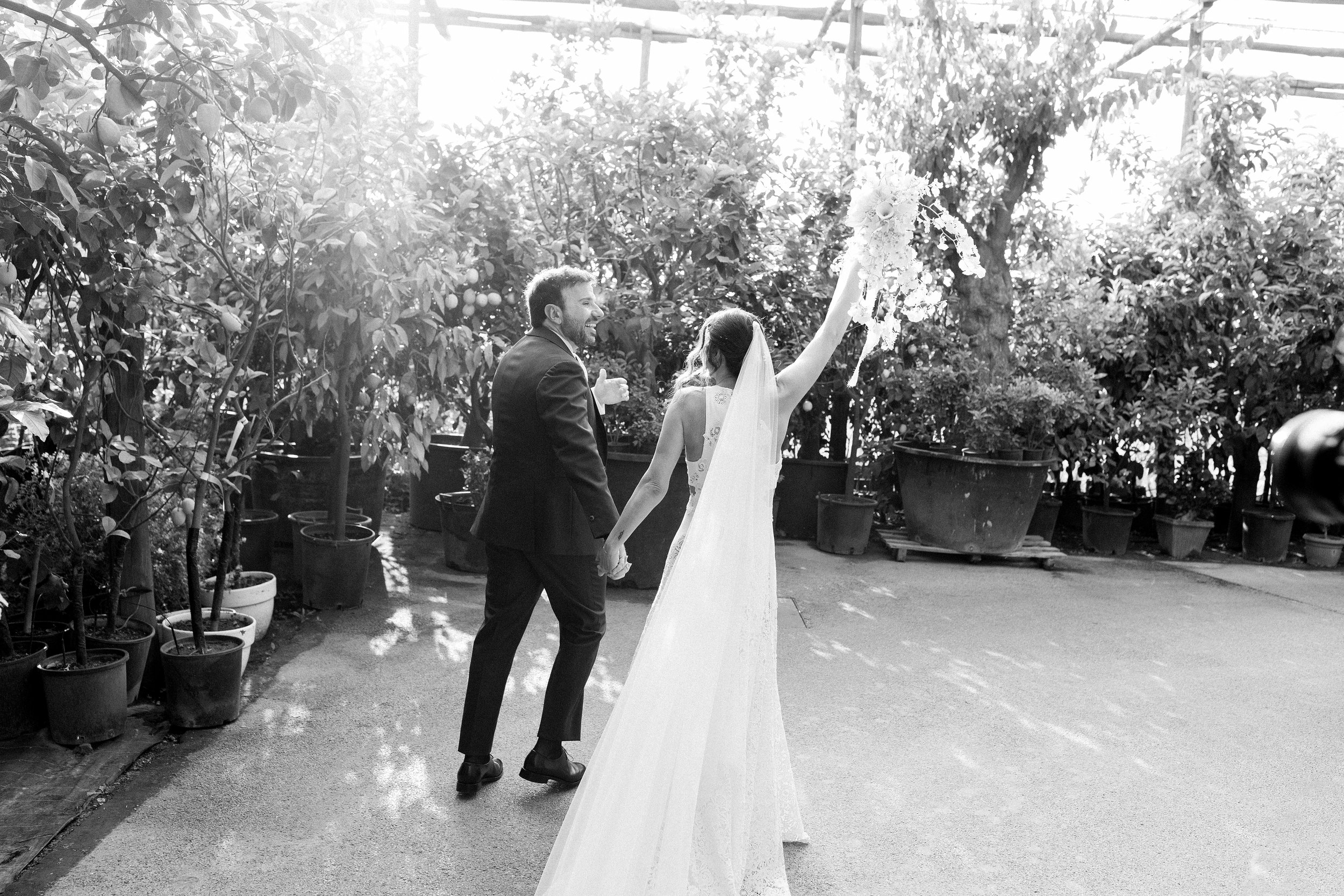 Una coppia di sposi felici si tiene per mano in un giardino, il/la sposa alza un bouquet di fiori, luci naturali creano un'atmosfera luminosa.