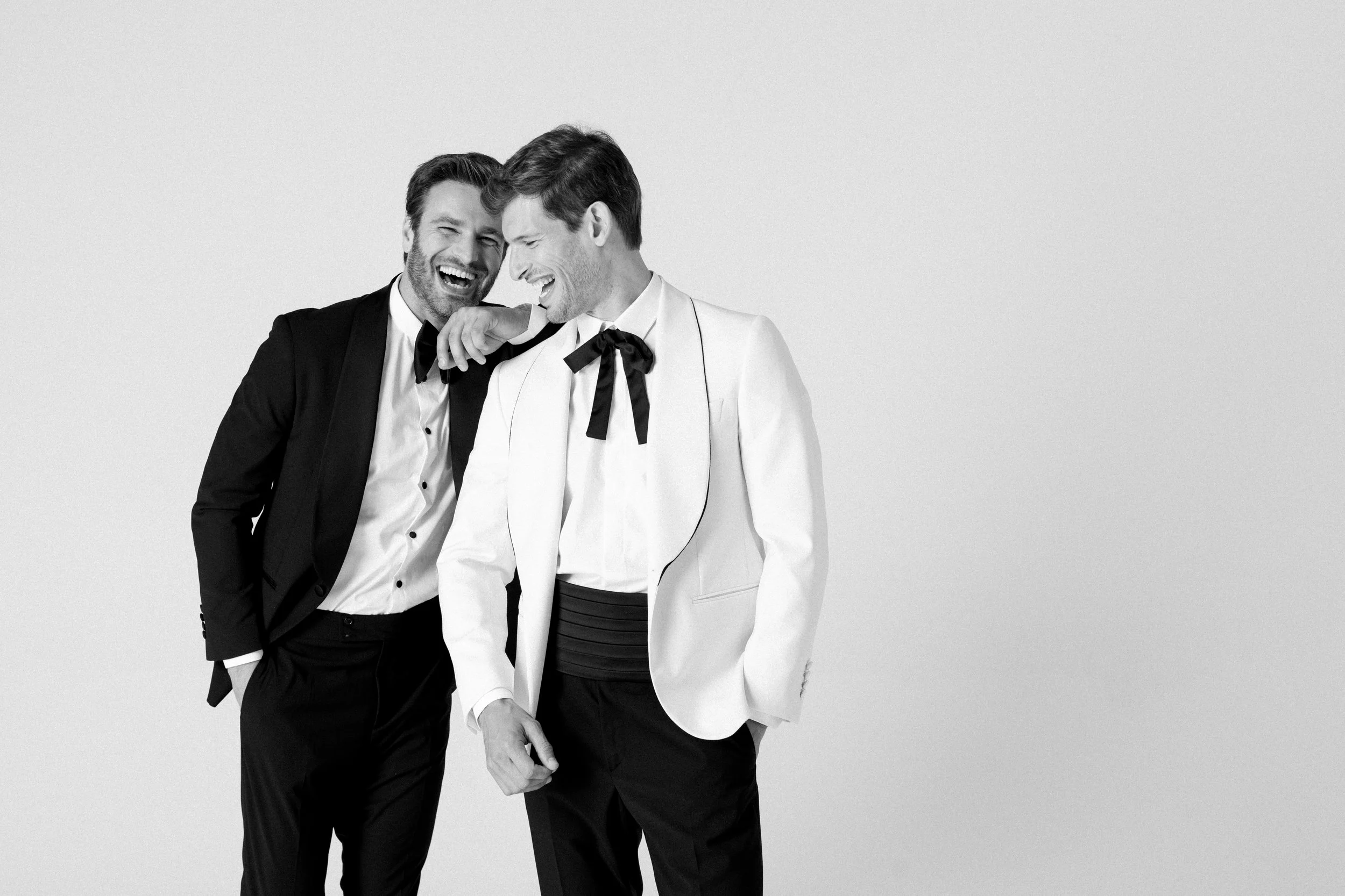 Due uomini sorridenti, magri e con capelli castani, indossano abiti eleganti e si divertono insieme. Uno indossa un abito nero con camicia bianca, l'altro indossa un abito bianco con sciarpa nera.