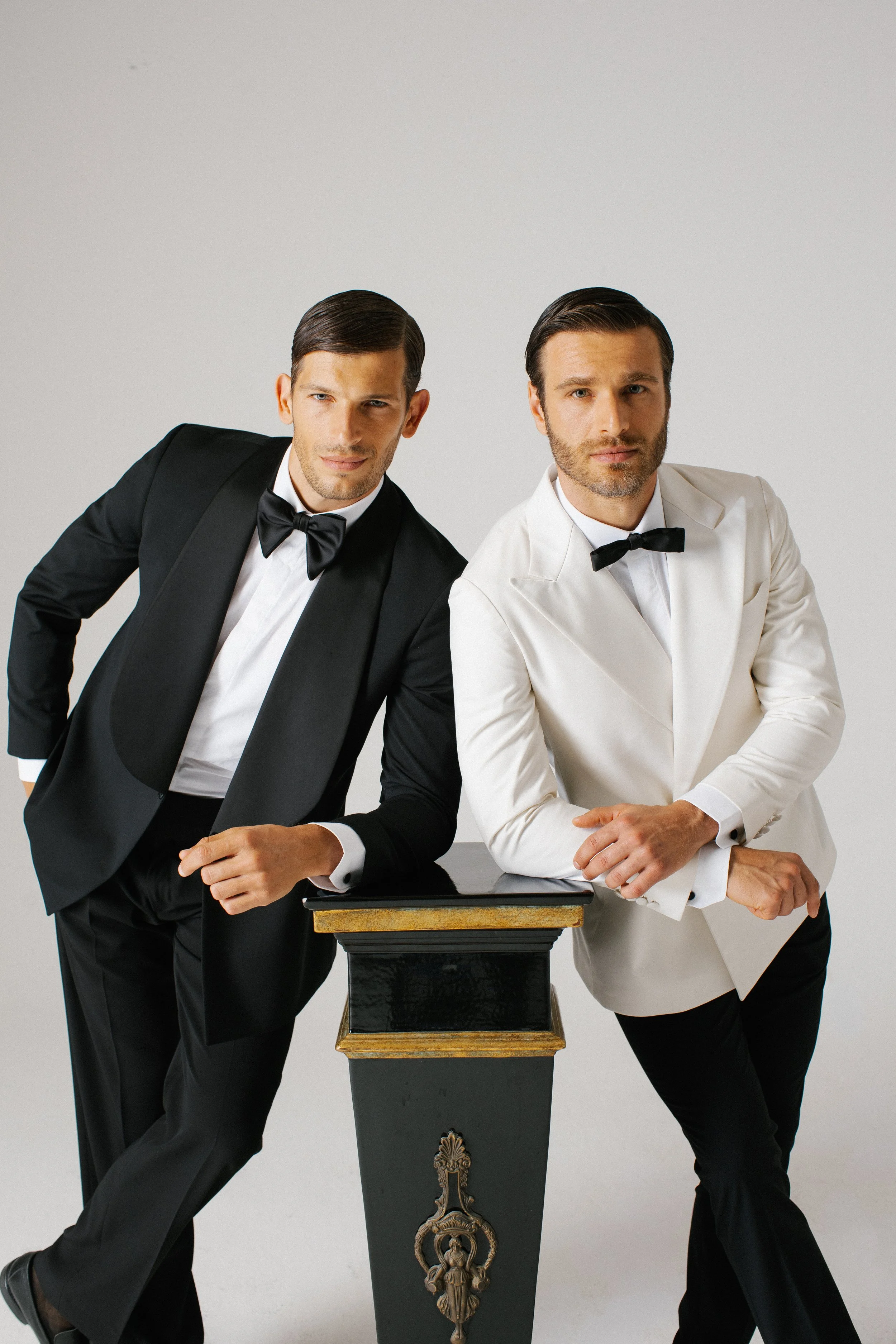 Due uomini eleganti vestiti con smoking, uno in nero e uno in bianco, posano con un piedistallo nero decorato.