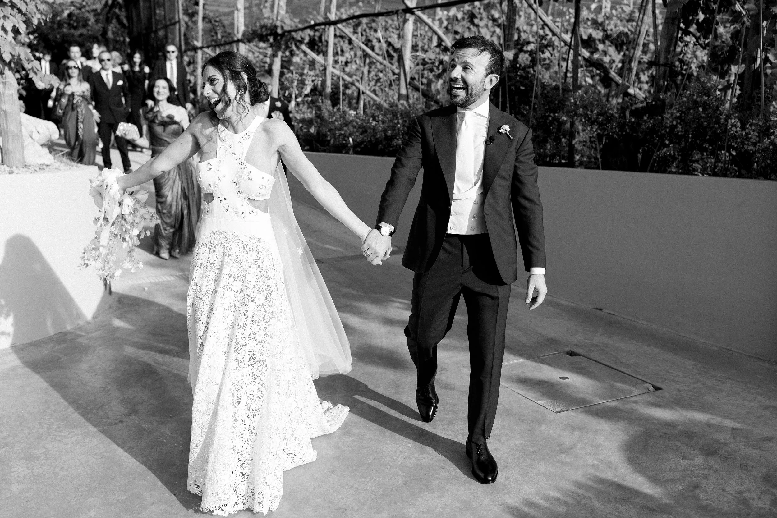 Una coppia di sposi, lei in abito da sposa e lui in abito elegante, tiene le mani mentre camminano sorridenti e felici, con amici e parenti sullo sfondo.