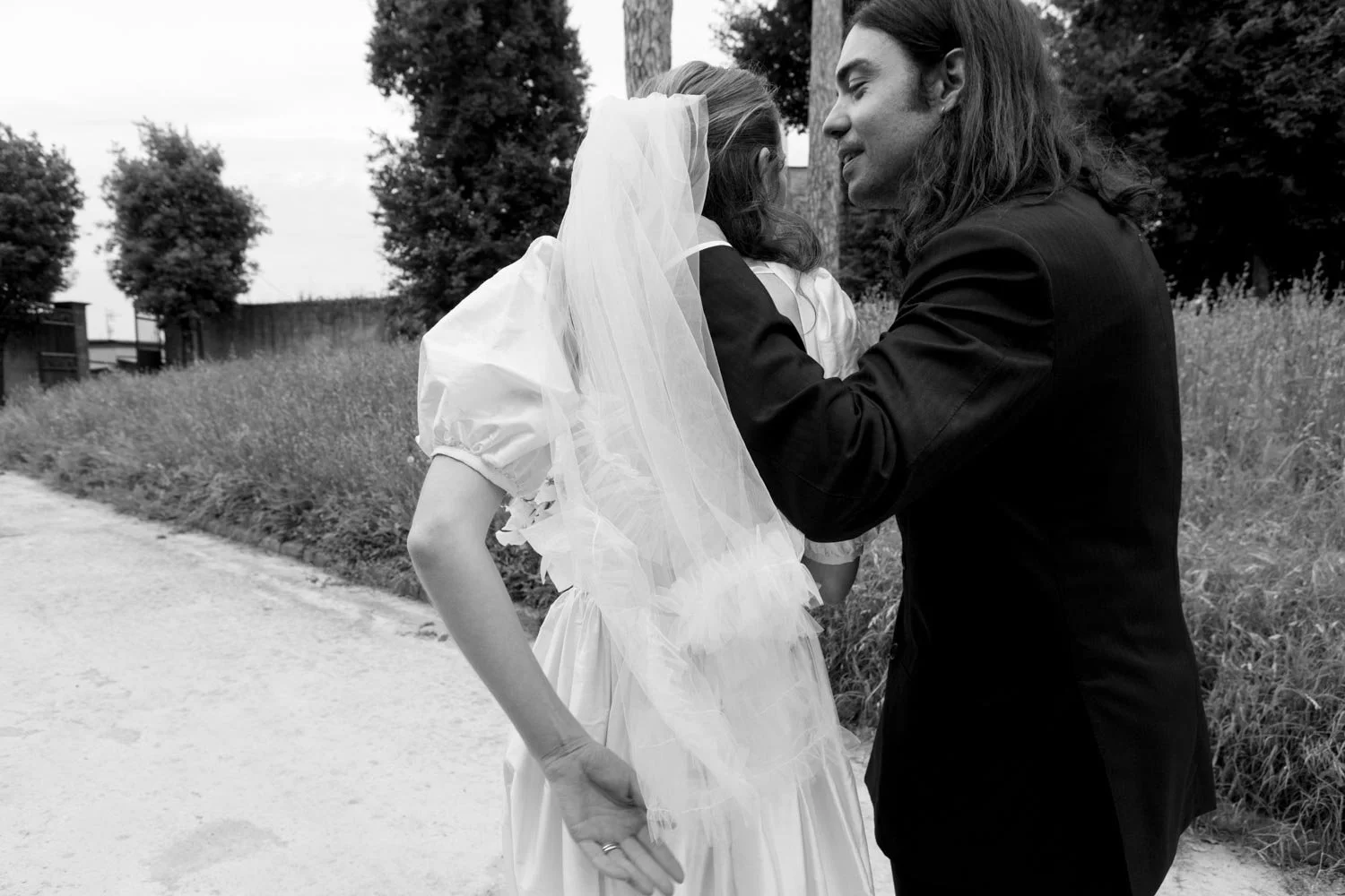 Una coppia di persone in abiti da matrimonio, con uno sfondo di natura, che si tengono per mano e si sorridono, in un’immagine in bianco e nero.