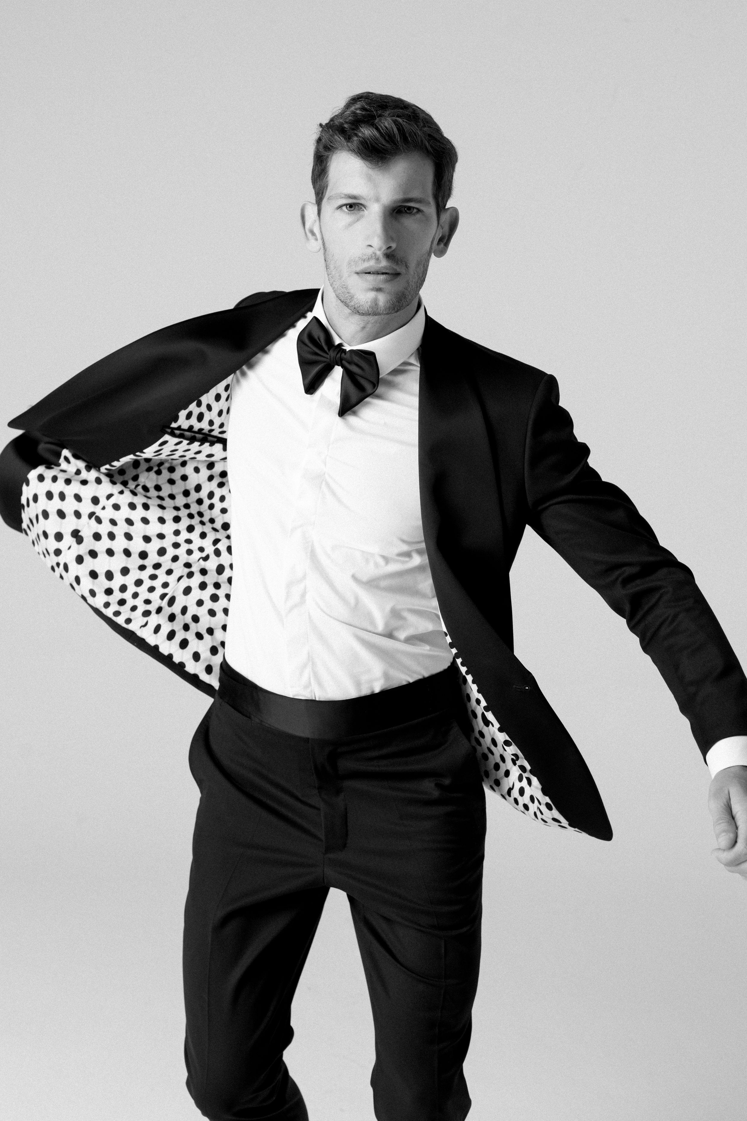 Uomo vestito con tuxedo nero, camicia bianca e fiocco nero, mentre si sistema la giacca, in un ambiente neutro, in bianco e nero.