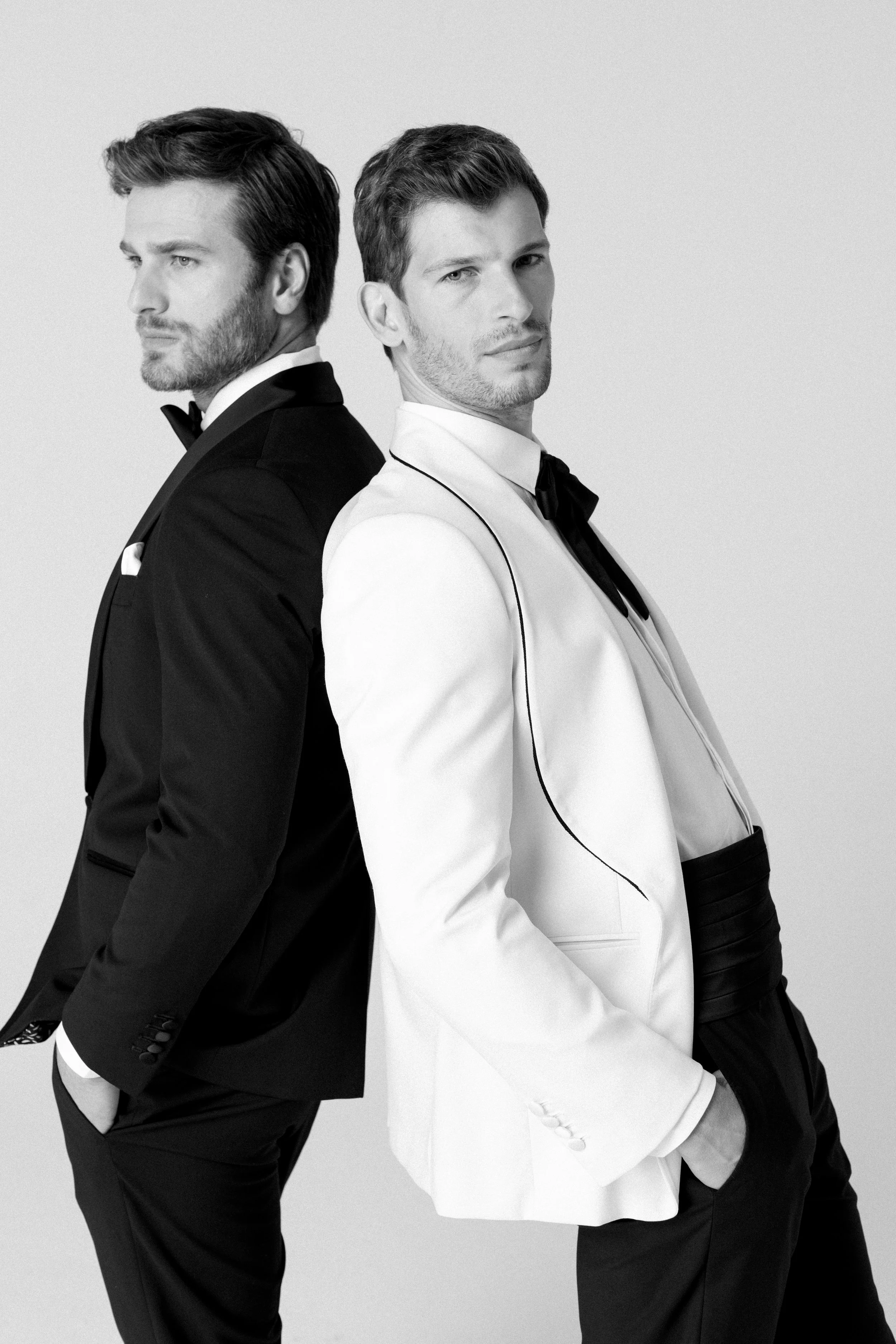 Due uomini in abiti da sera, uno in tuxedo nero e l'altro in smoking bianco, che si sdraiano con le schiene l'uno contro l'altro, in posa su sfondo grigio chiaro.