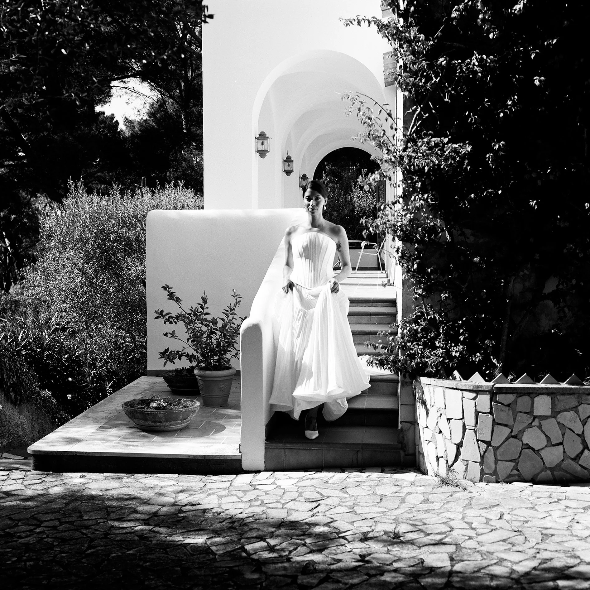 Donna in abito da sposa che scende le scale in un ambiente all'aperto con piante e strutture architettoniche in bianco e nero.