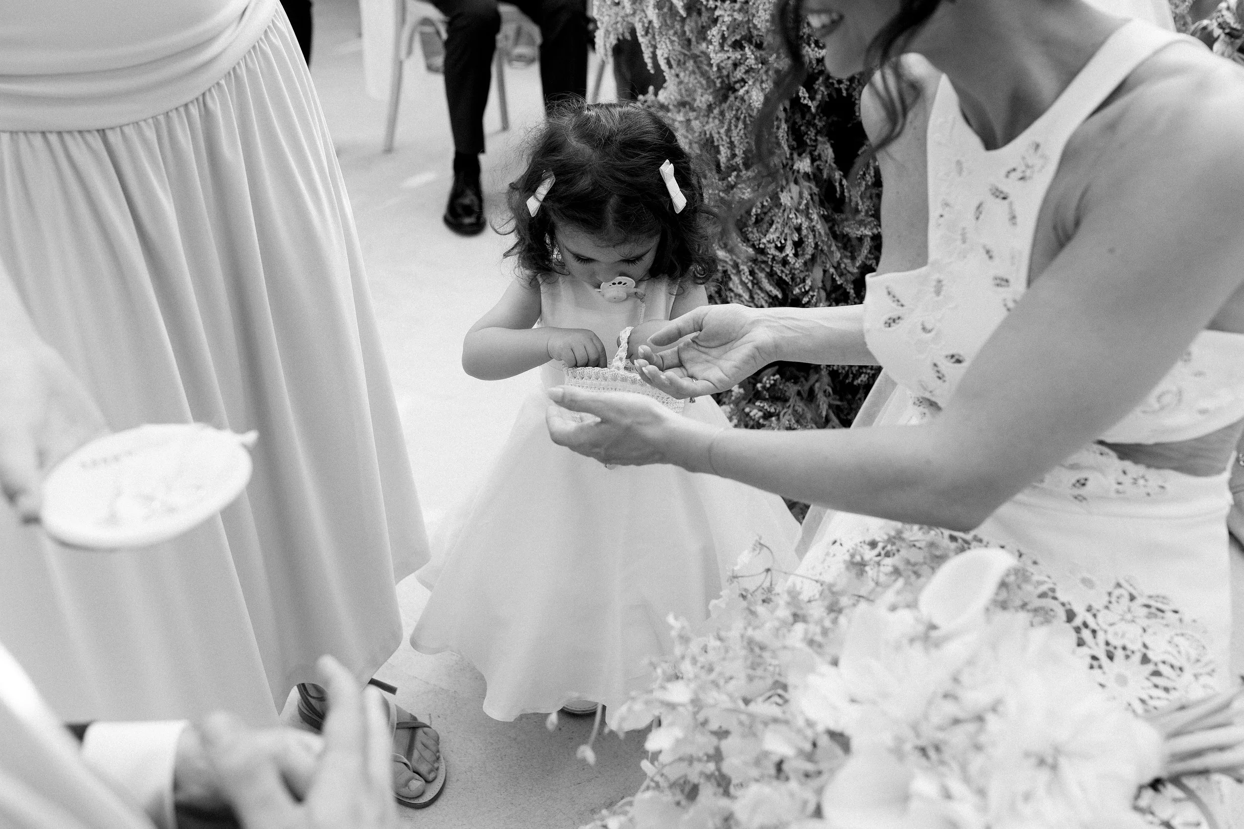 Una bambina con un biberon in bocca che riceve un chicco di riso da una donna in un contesto di festa. La scena è standosa e intima, probabilmente durante un ricevimento di matrimonio o una celebrazione simile.