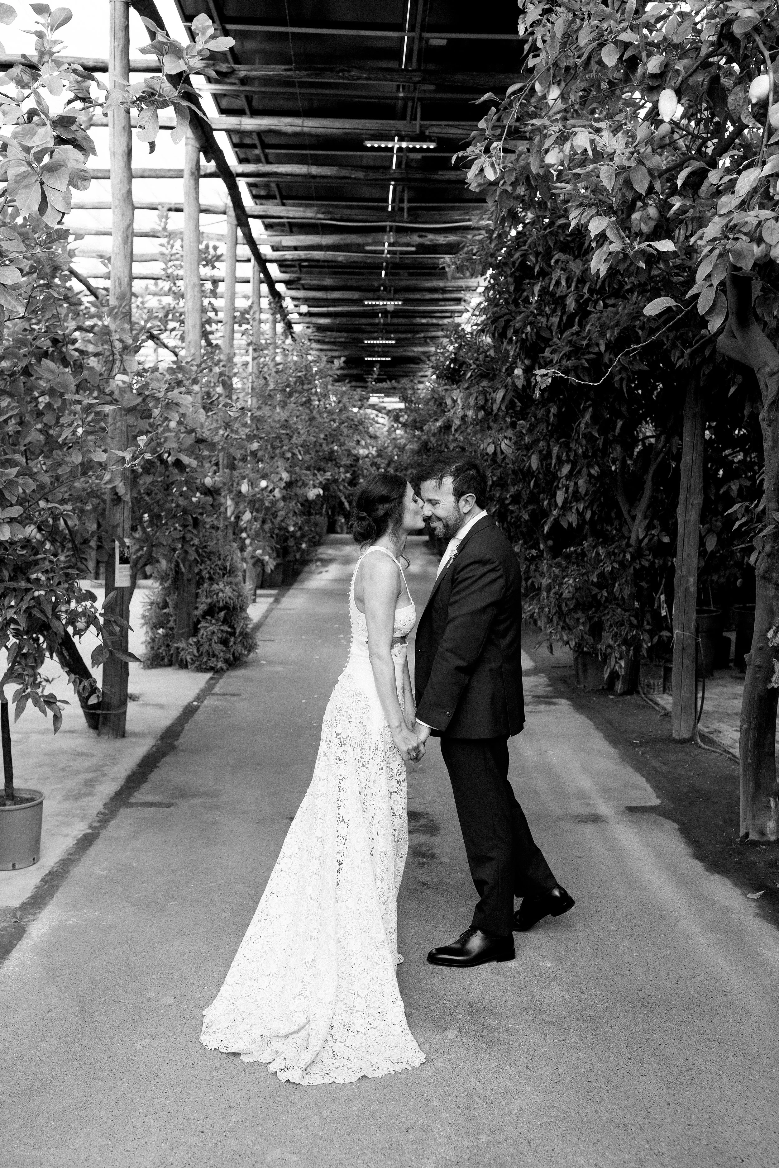 Una coppia di sposi si bacia nel centro di un vialetto in un giardino o serra, circondati da piante e alberi.