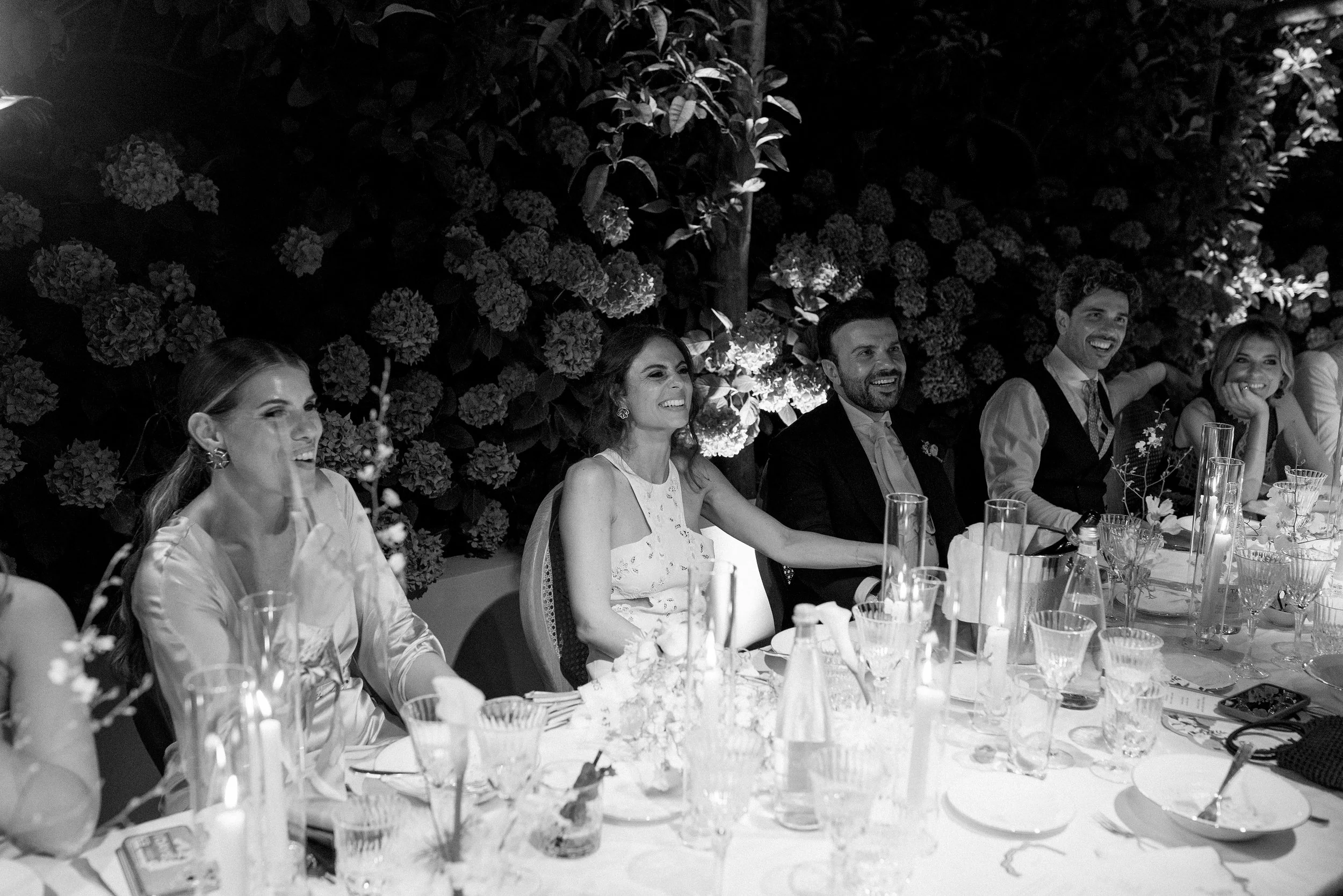 Gente seduta a un tavolo durante una festa, sorridendo e divertendosi, con decorazioni floreali sullo sfondo.