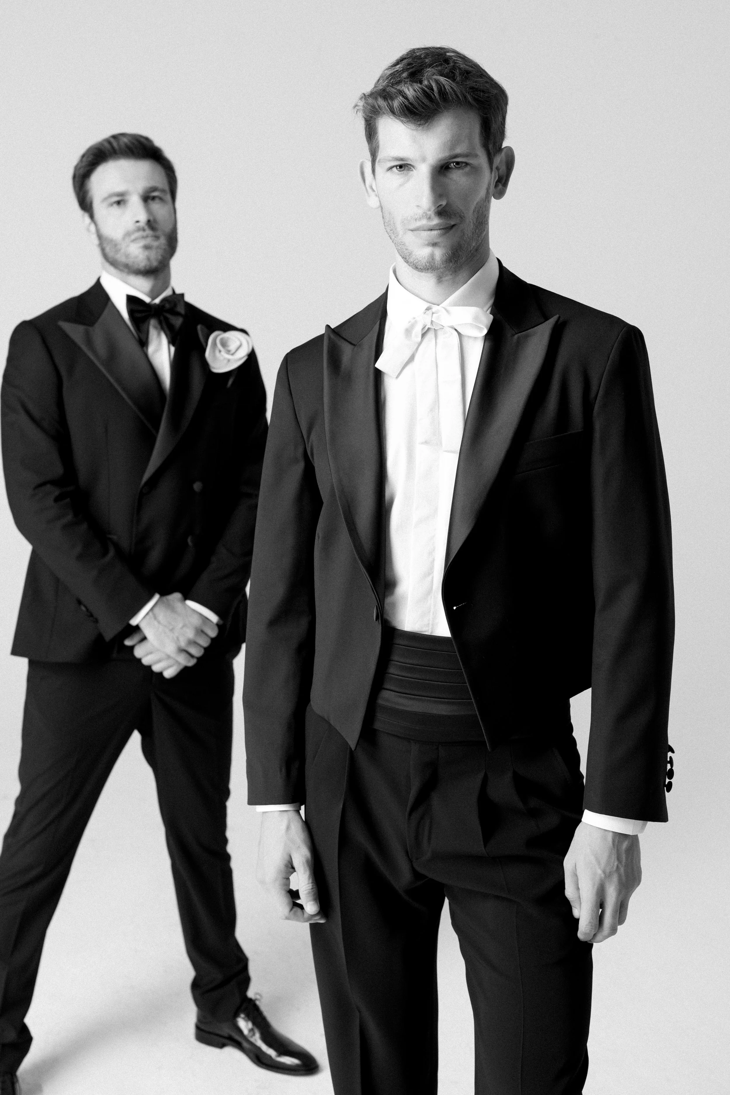 Due uomini indossano abiti eleganti da sera, uno in primo piano e l'altro sullo sfondo, entrambi con abiti neri e cravatte bianche, in un'immagine in bianco e nero.
