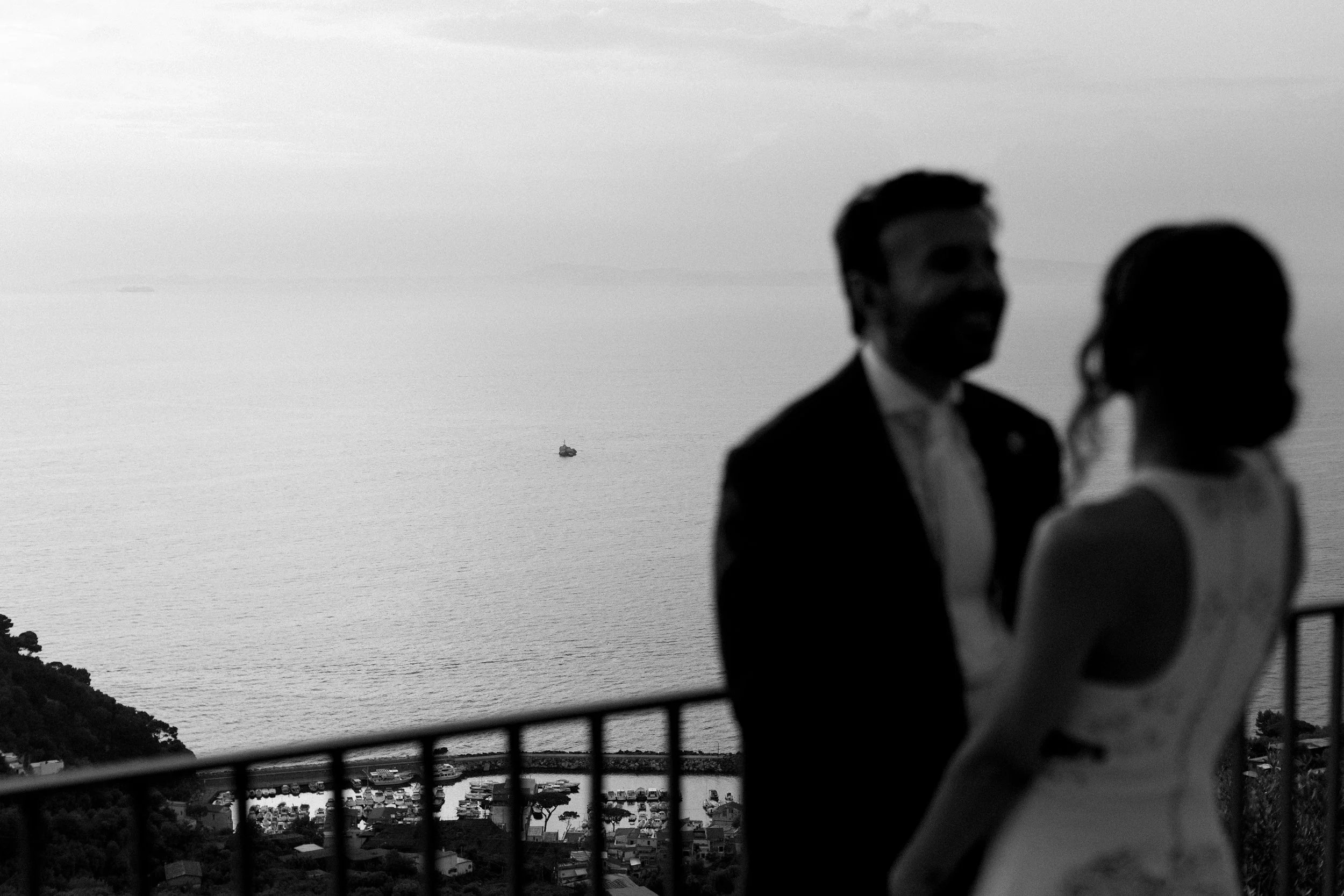 Due persone, un uomo e una donna, in abiti eleganti si abbracciano su un balcone con vista sul mare, con una barca visibile in lontananza.