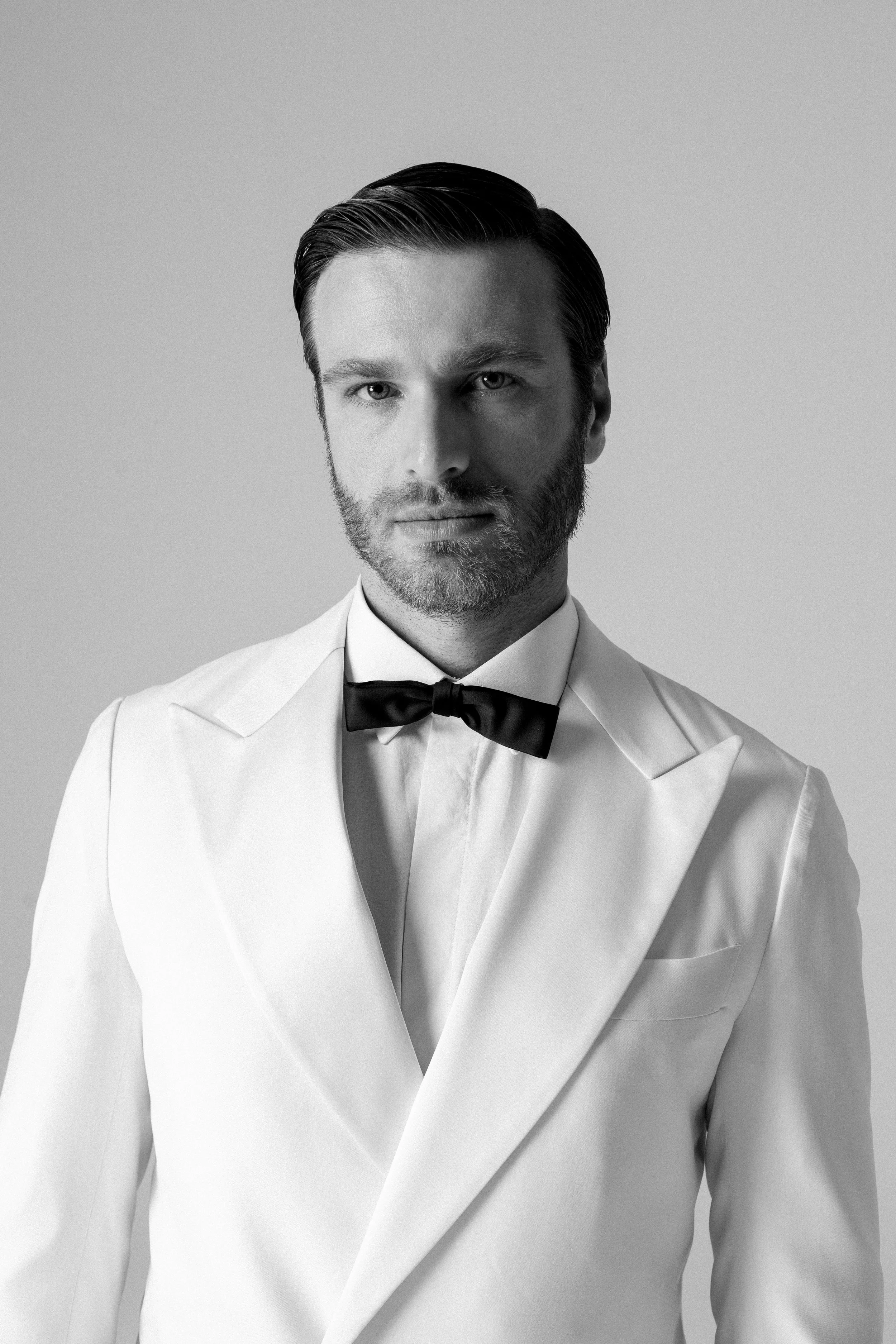 Uomo con barba e cappello in bianco e nero, vestito elegante con giacca bianca e papillon nero.