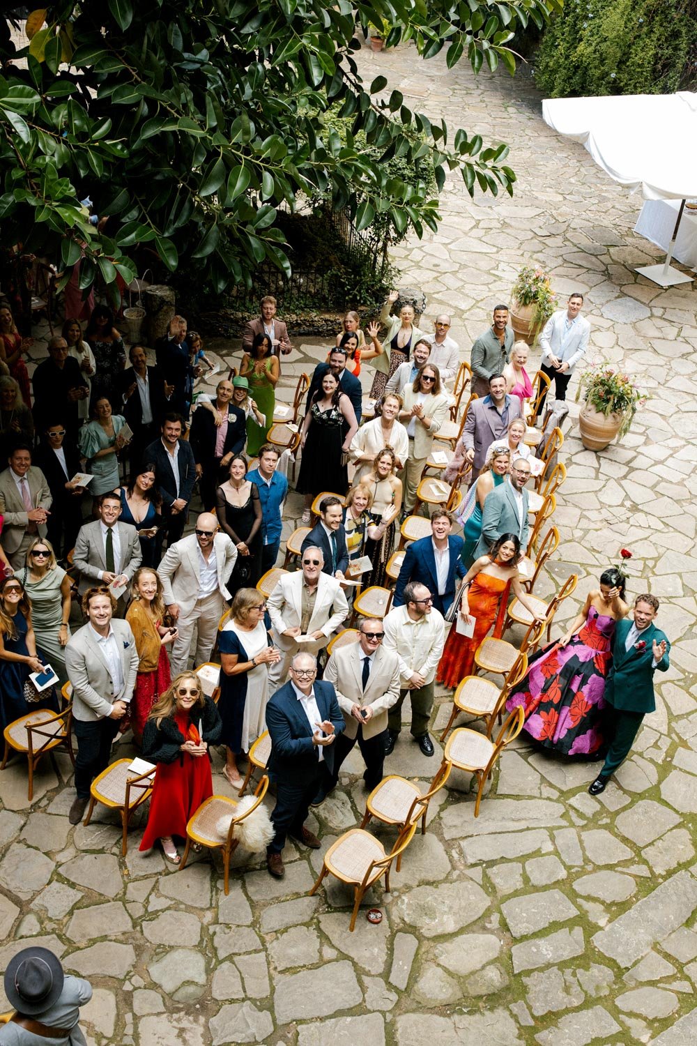 Un gruppo di persone sorridenti e ben vestite in un cortile all'aperto, alcune sedute su sedie di legno, in occasione di un evento sociale o matrimonio, con piante e una copertura bianca sullo sfondo.