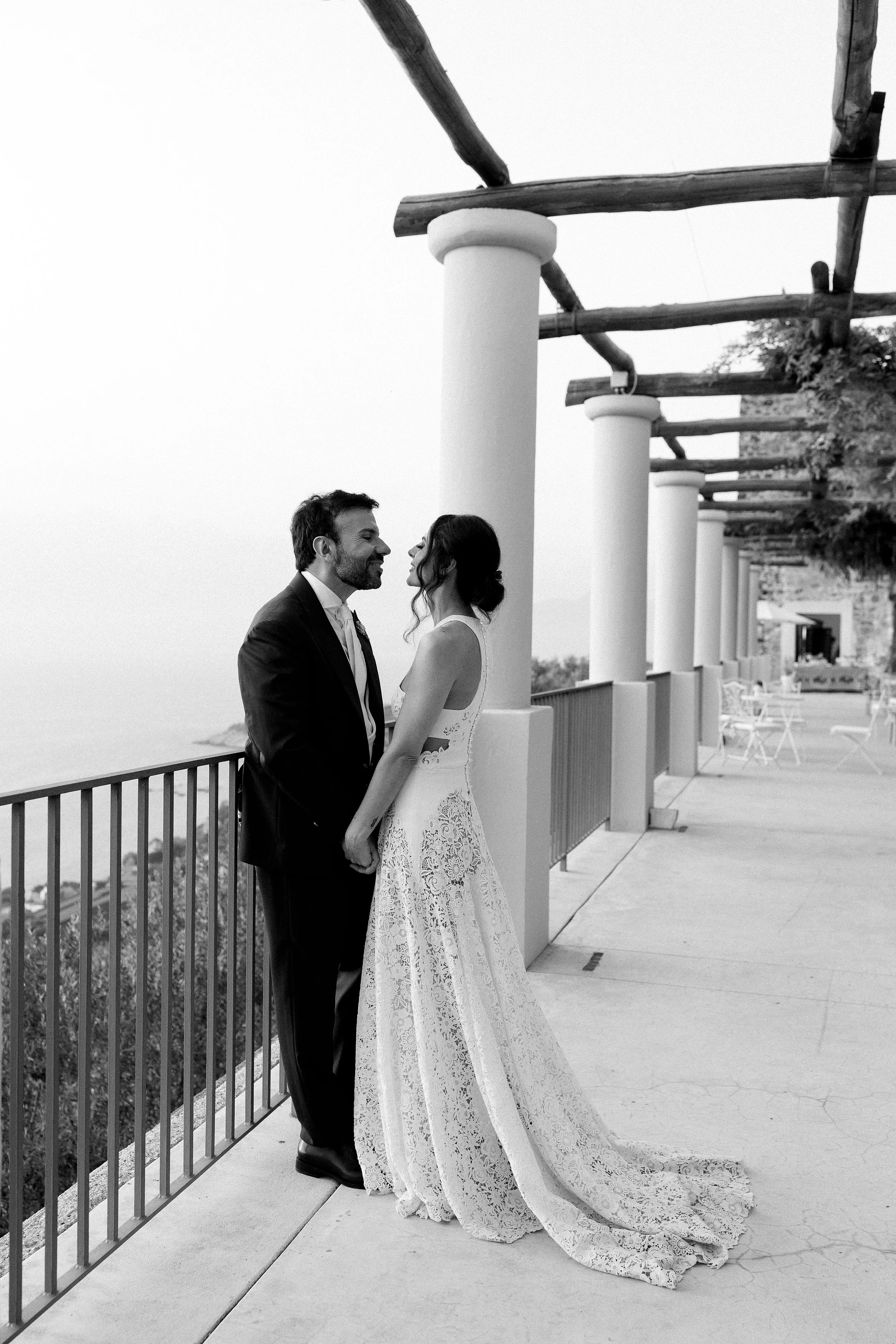 Una coppia di sposi in abiti da matrimonio si guarda intensamente su una terrazza con colonne, in bianco e nero.