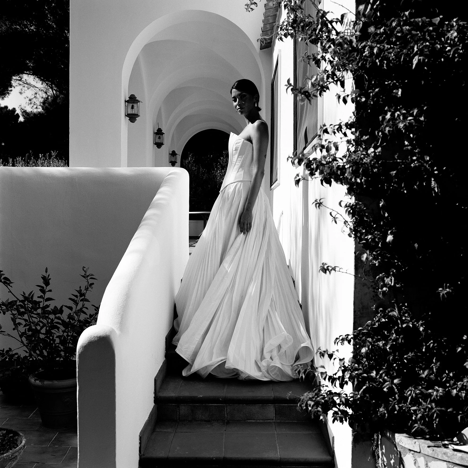 Donna in un abito da sposa bianco su una scala, con archi e luci all'esterno, in una scena in bianco e nero.