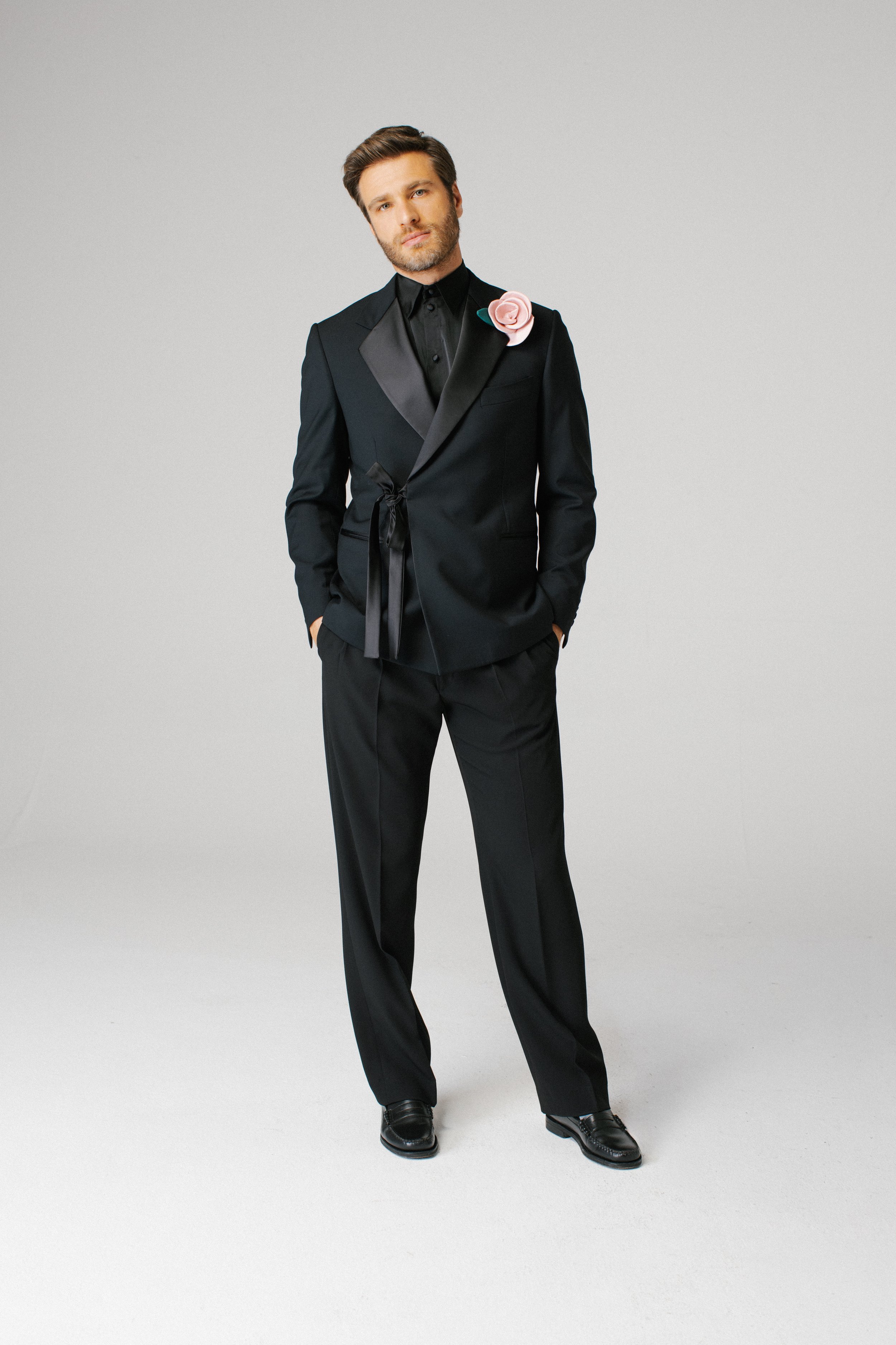 Uomo in completo nero con fiocco rosa a fianco, su sfondo grigio chiaro.