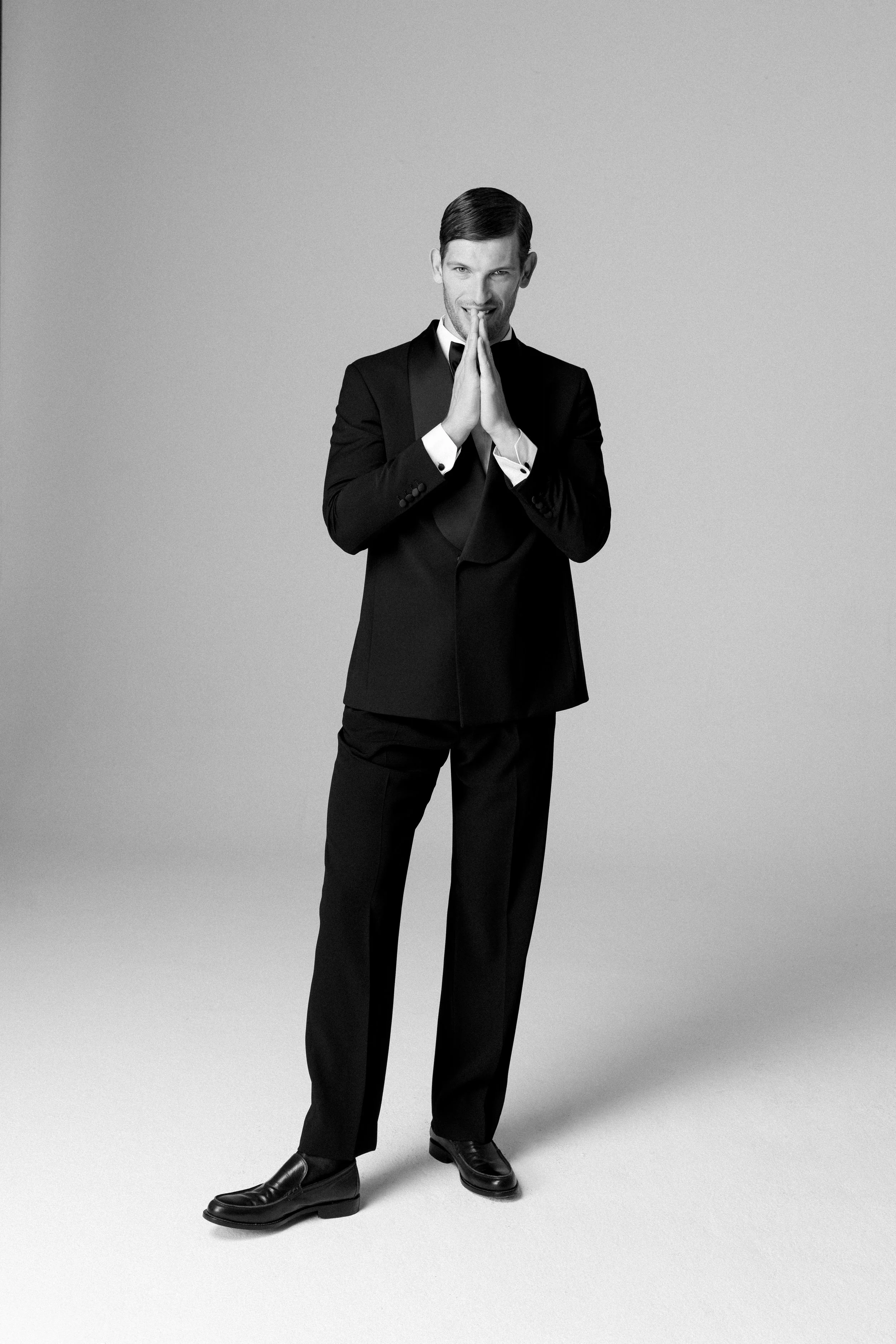 Uomo in abito nero, in posa con le mani giunte davanti al viso, sorriso, sfondo grigio.