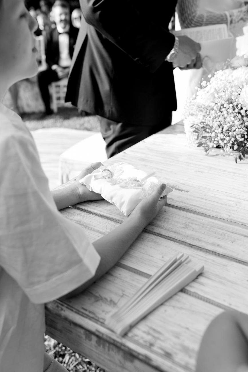 Bambina con manette di matrimonio in mano, seduta su un tavolo di legno, con anelli nuziali su un cuscino, in un matrimonio all'aperto. Sul tavolo ci sono anche alcune cartoline o biglietti.
