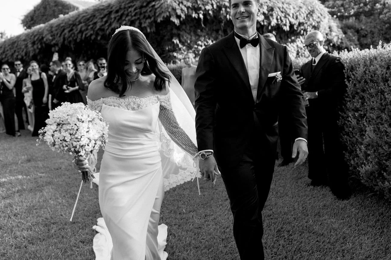 Coppia di sposi che camminano mano nella mano durante il matrimonio, con invitati sullo sfondo, in un giardino.